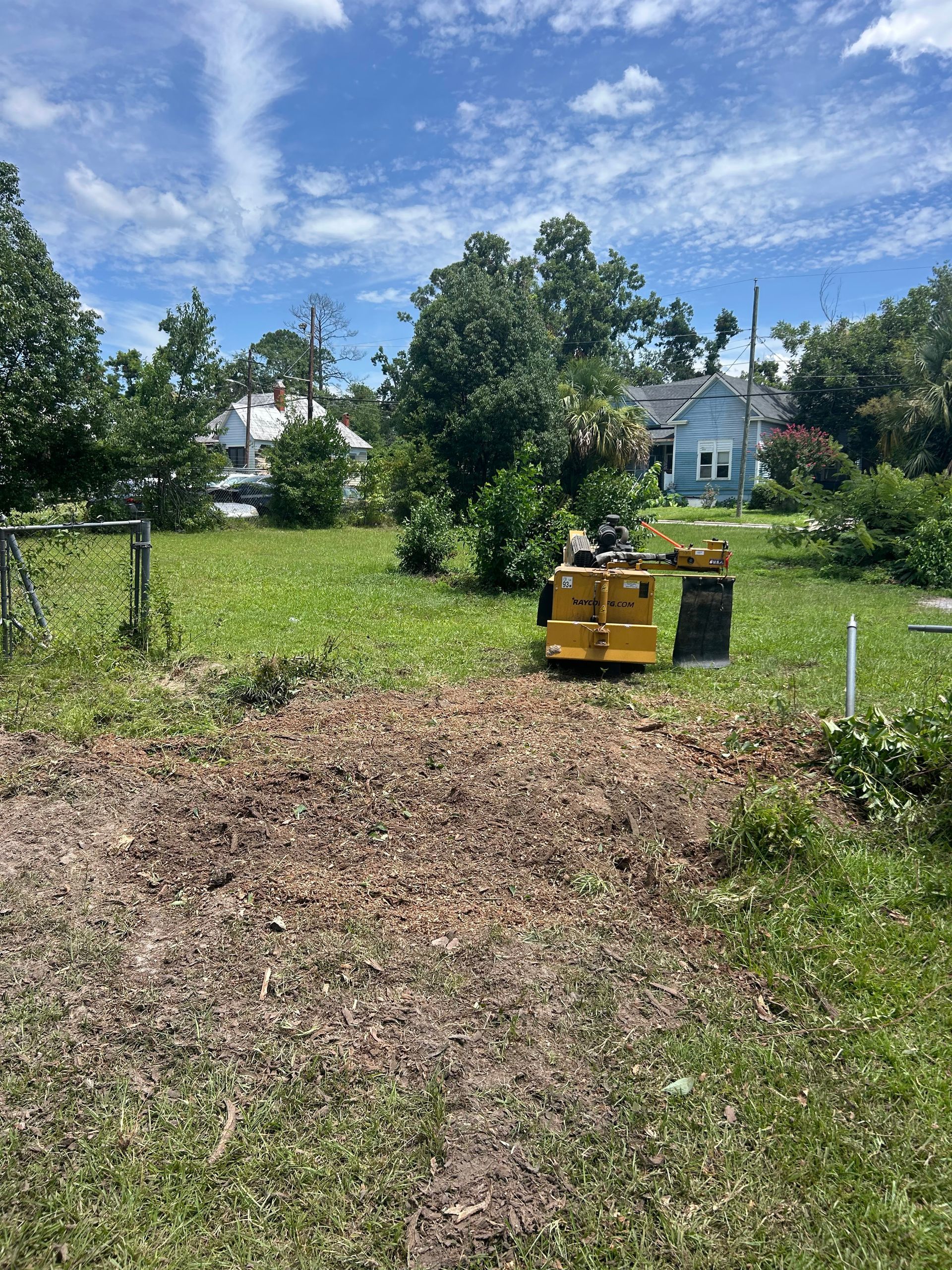 Grassy yard with trees, houses, and a yellow-green machine on the right under a blue sky.