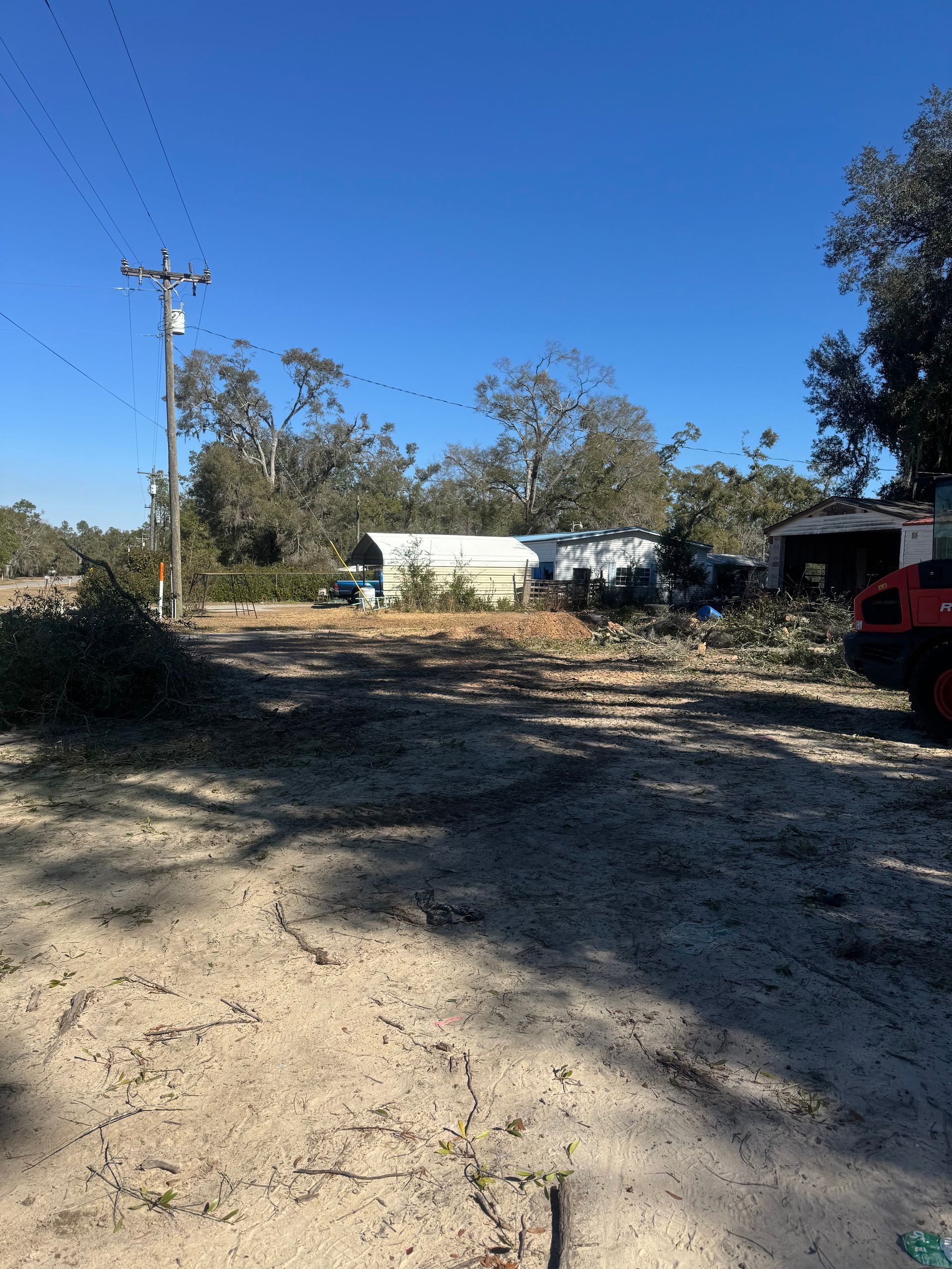 Rural dirt driveway with trees, a white building, utility pole, and a parked red vehicle under a clear blue sky