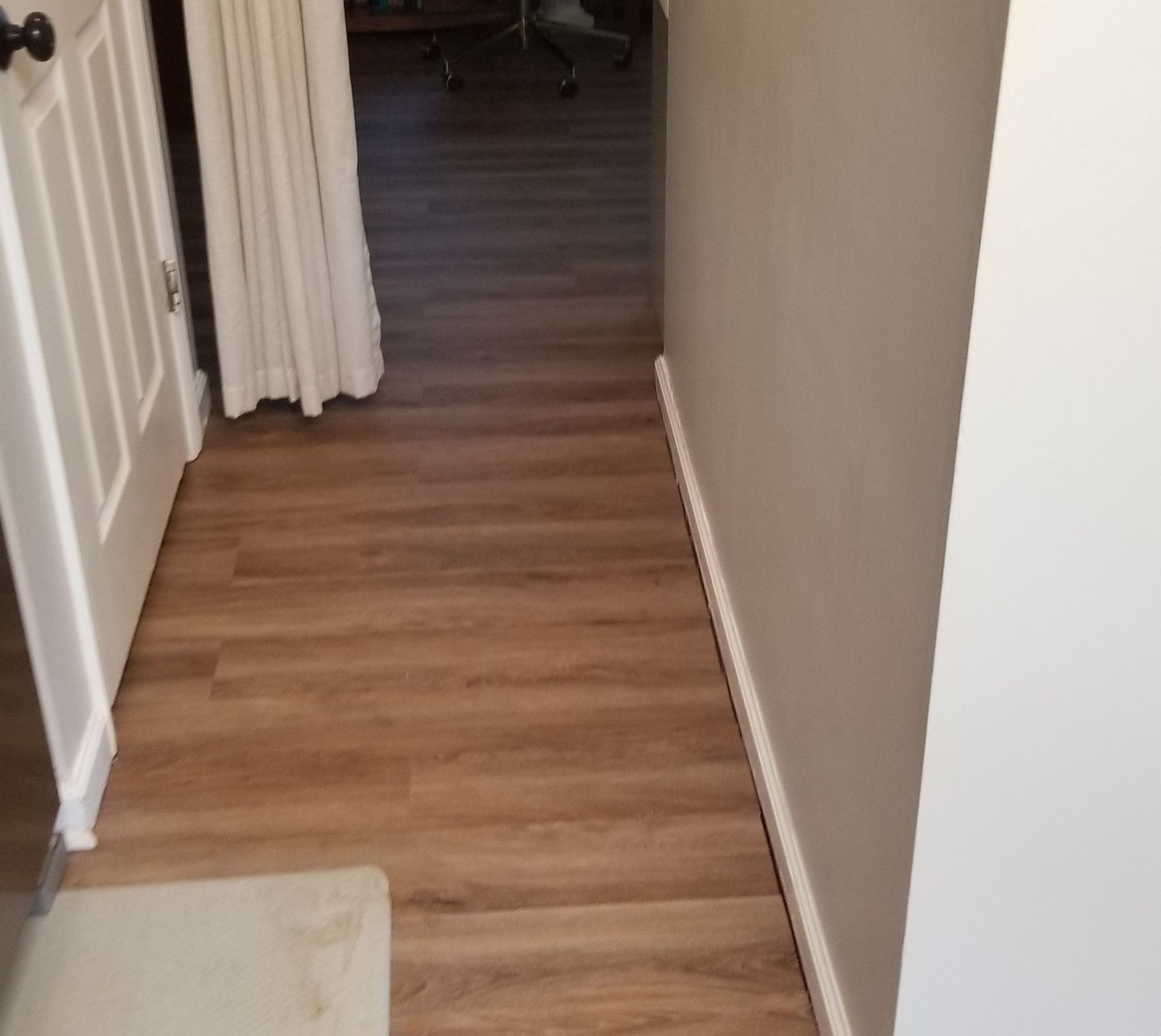A hallway with light brown wood-look flooring, a white door on the left, a curtained doorway, and light gray walls.