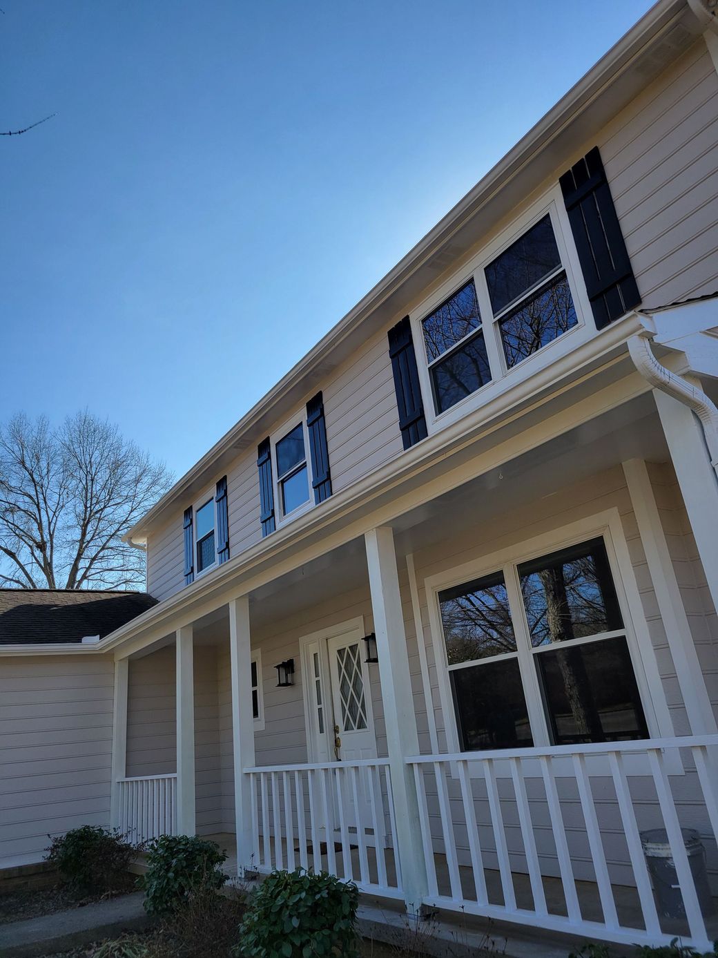 A low-angle view of a beige, two-story house with black shutters, a white front porch railing, and a clear blue sky.