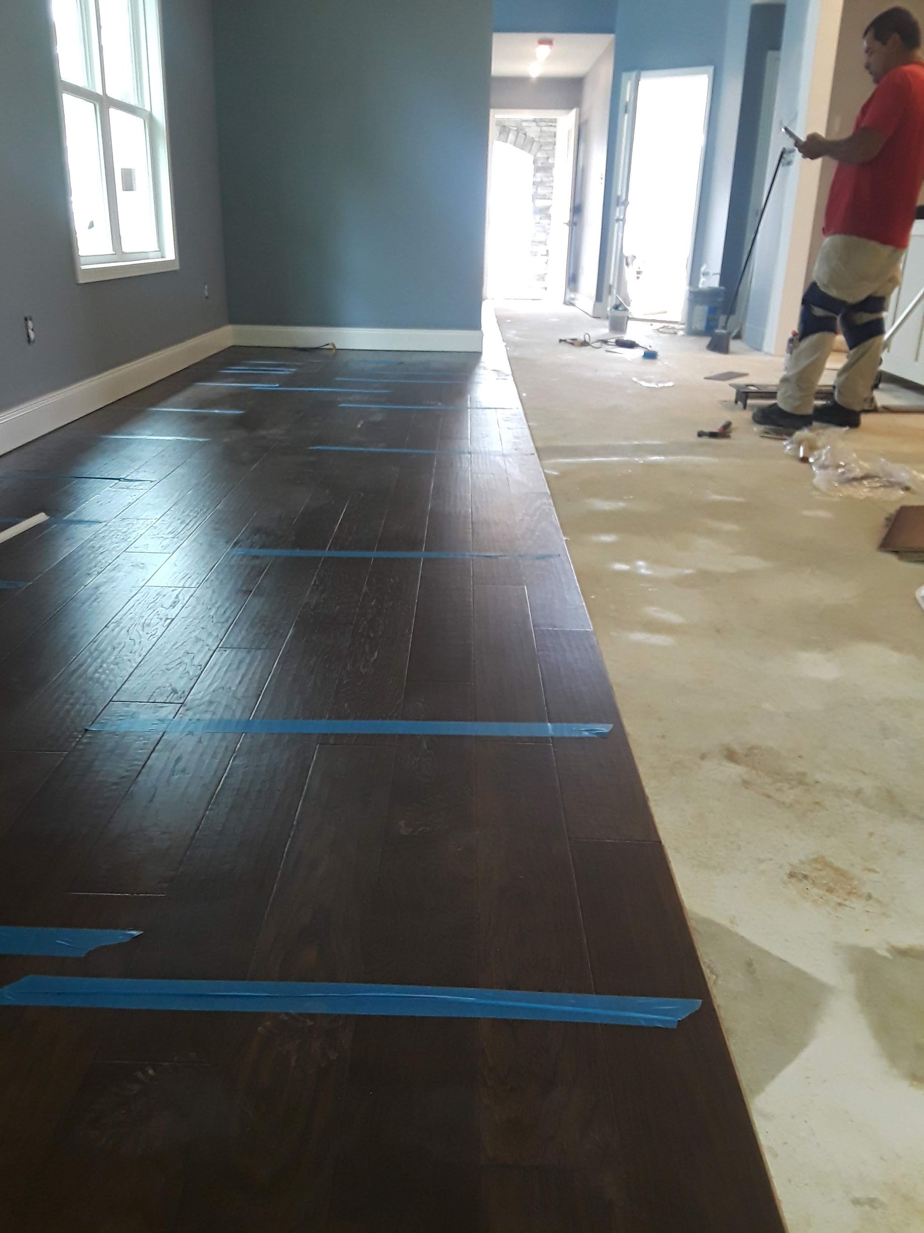 A person in a red shirt works in a room with partially installed dark wood flooring and blue painter's tape markings.