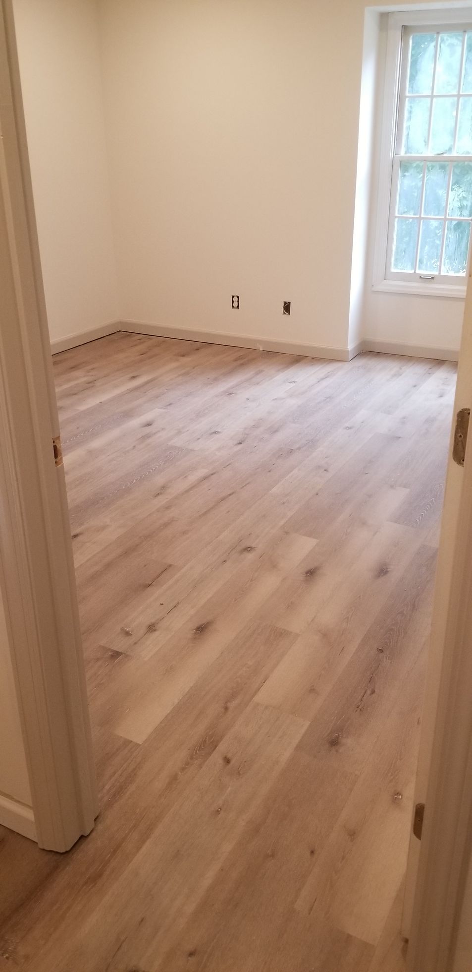 An interior view of a room featuring light-colored hardwood flooring, cream-colored walls, and a window with natural light.