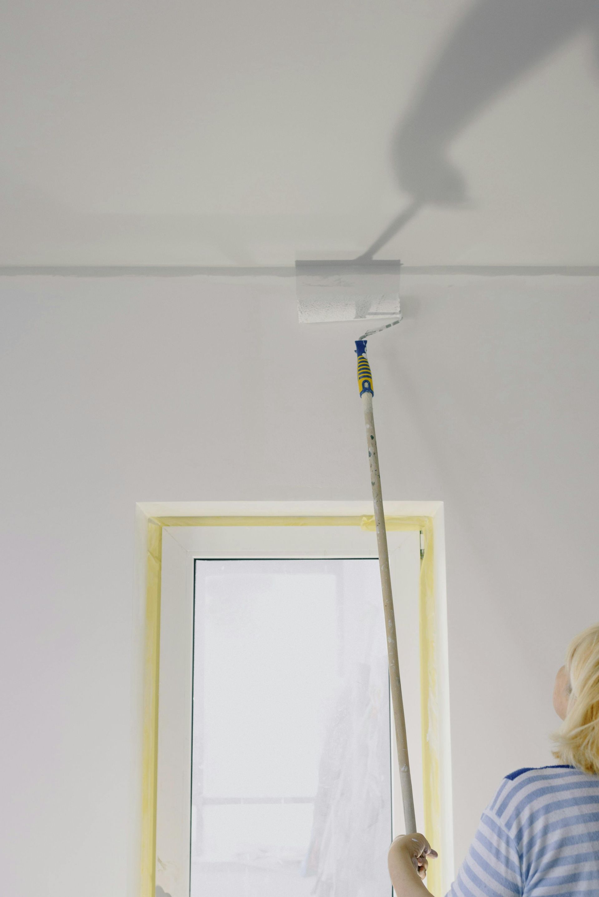 A person uses a paint roller on an extension pole to paint a white ceiling near a window.