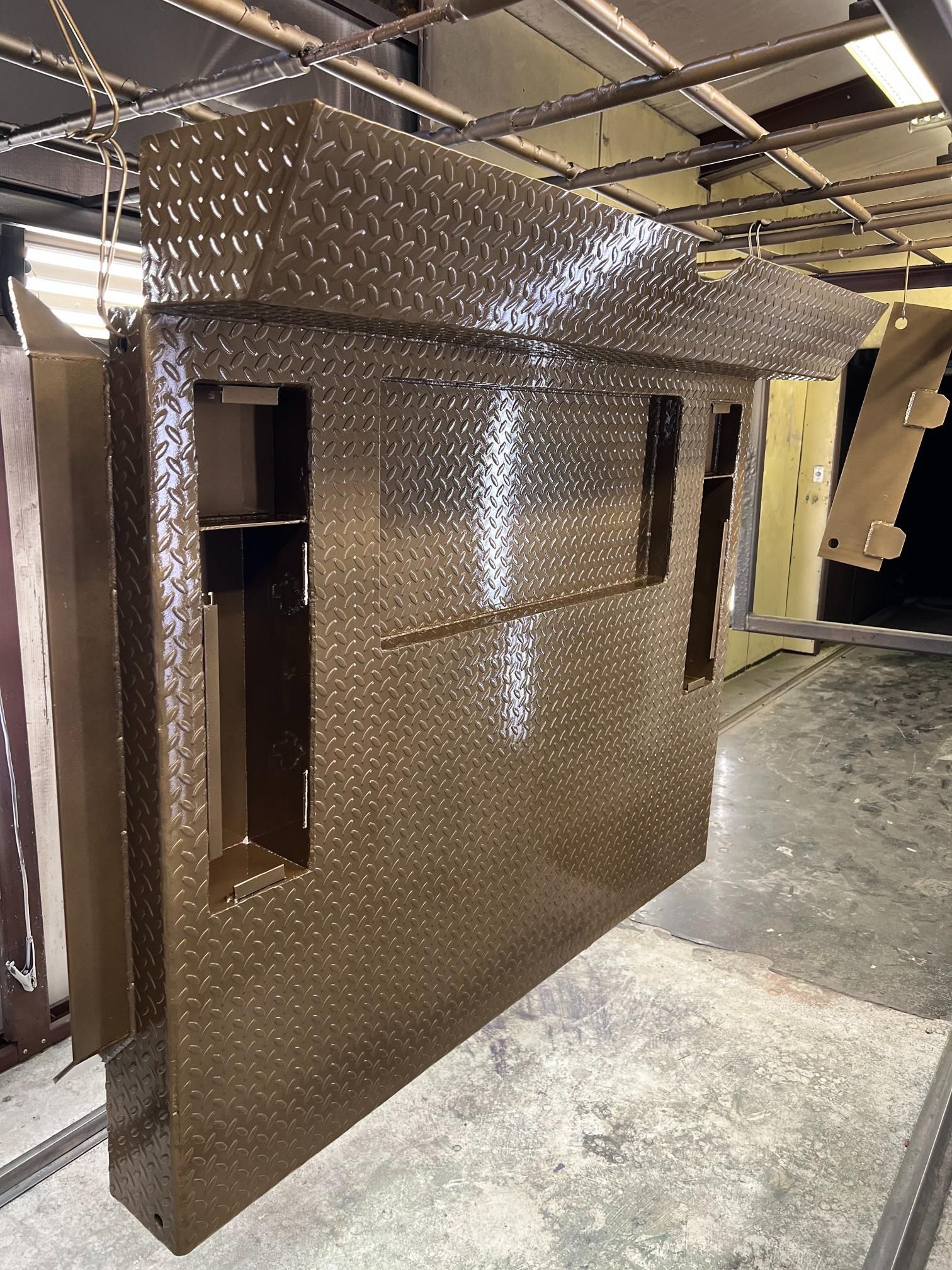 A brown diamond-plate metal panel with cutouts hangs from an overhead rack in a warehouse-style setting.
