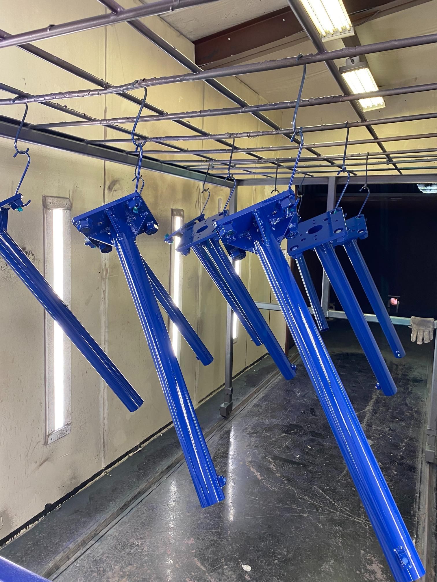 Several bright blue metal components hang from hooks in an industrial spray booth for powder coating.
