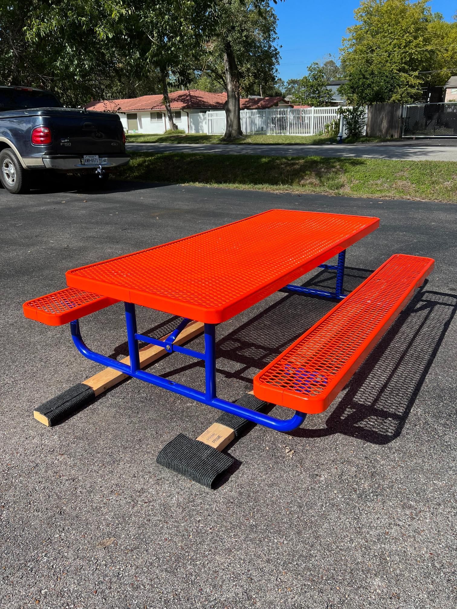 A bright orange, metal picnic table with a blue frame stands on wooden blocks in a paved parking lot.