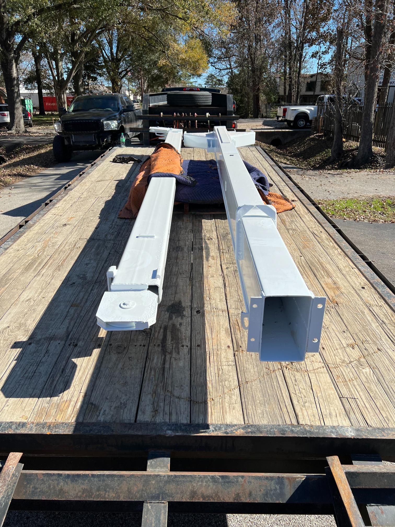 Two long, white metal support columns lie on a flatbed trailer outdoors, with a truck visible in the background.