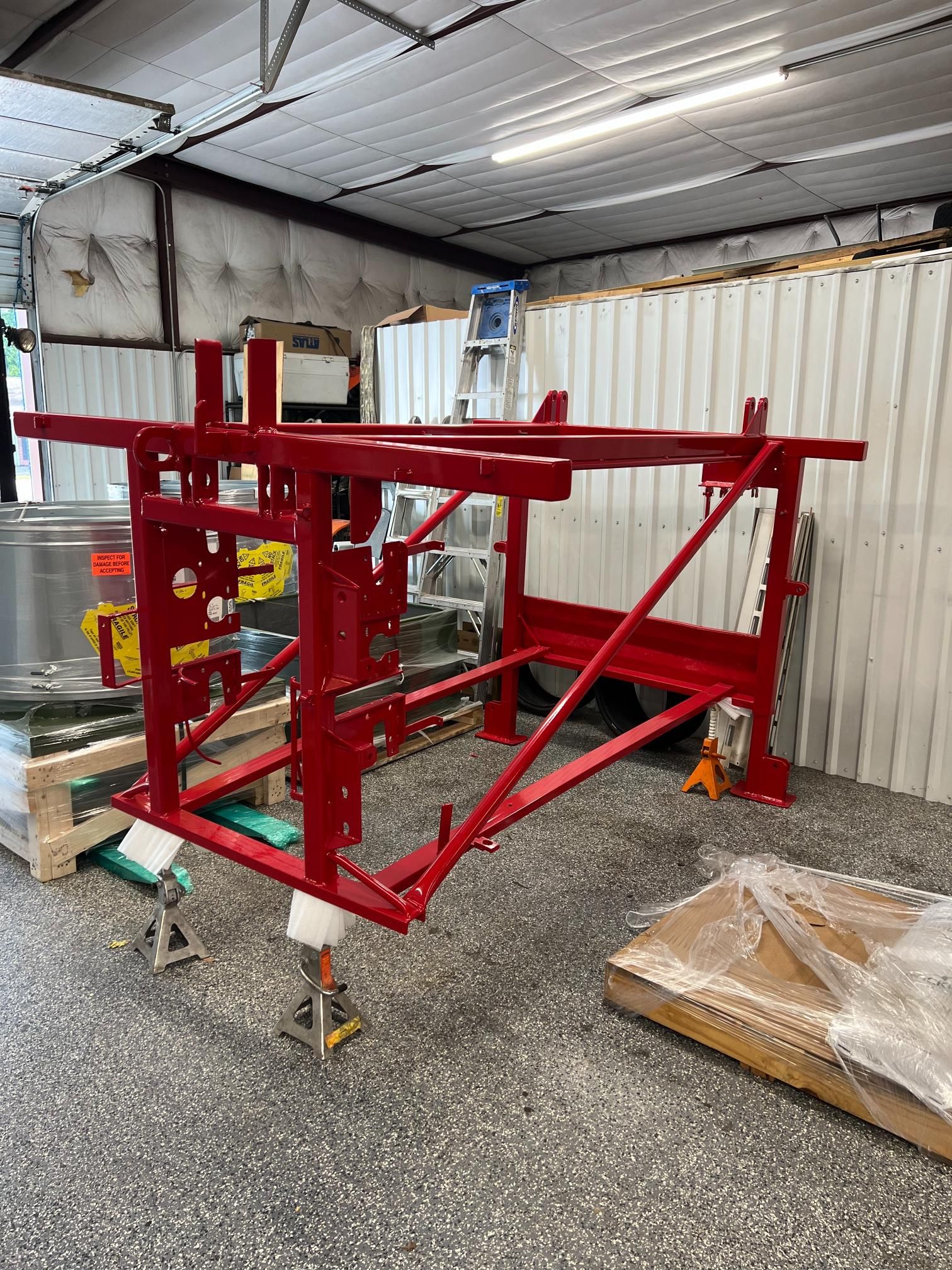 A red, boxy metal frame structure stands on jack stands in a workshop with a corrugated metal wall.