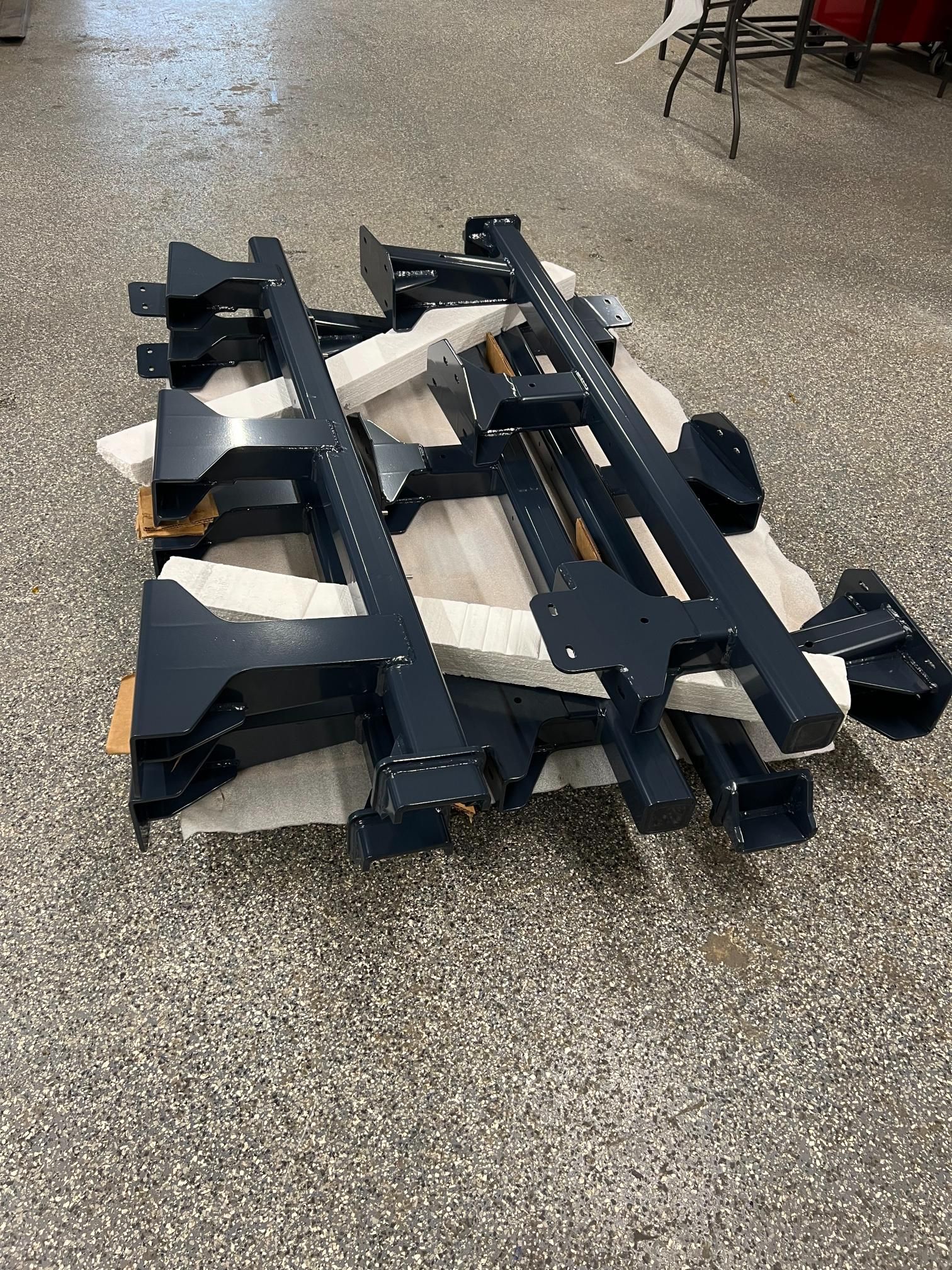 A stack of black steel structural frames or pallet rack components resting on a wooden pallet on a concrete floor.