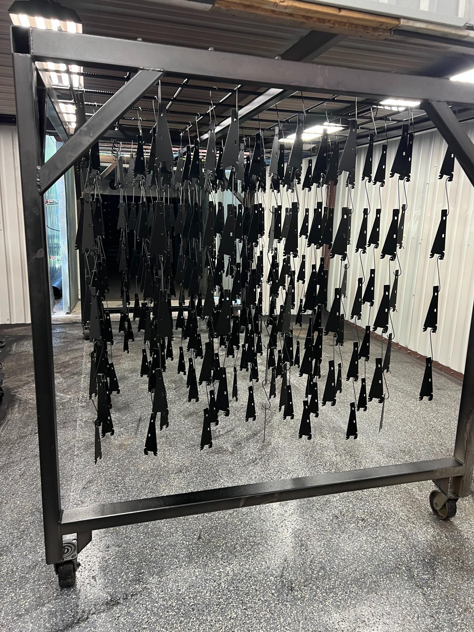 A black metal rack holds dozens of identical black components hanging from wires in an industrial workshop.