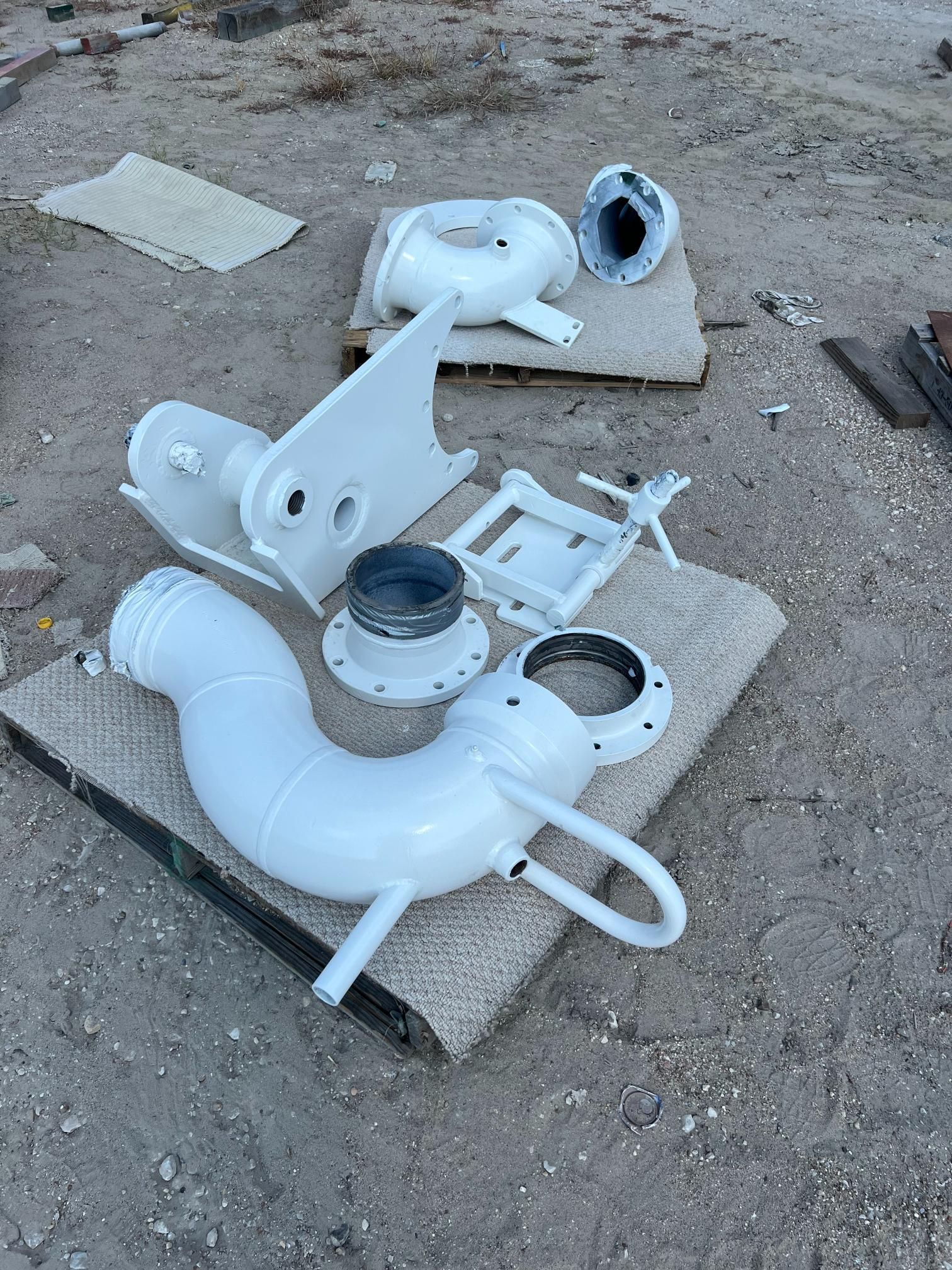 White painted metal industrial pipe fittings and a structural bracket resting on pieces of carpet outdoors on the ground.