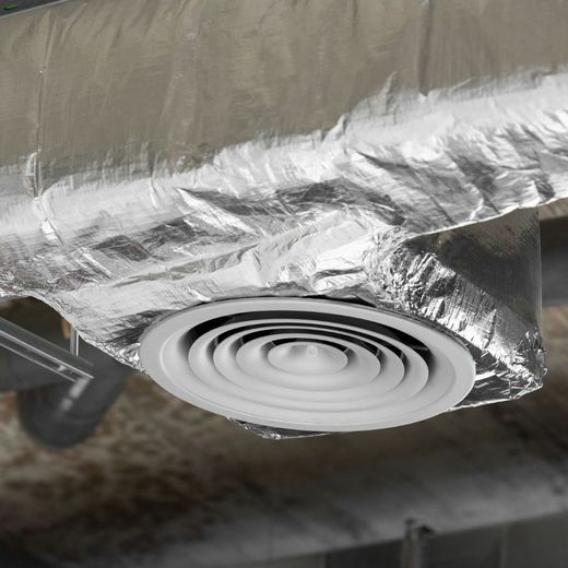 A white, circular HVAC ceiling diffuser installed in a silver, foil-insulated air duct.