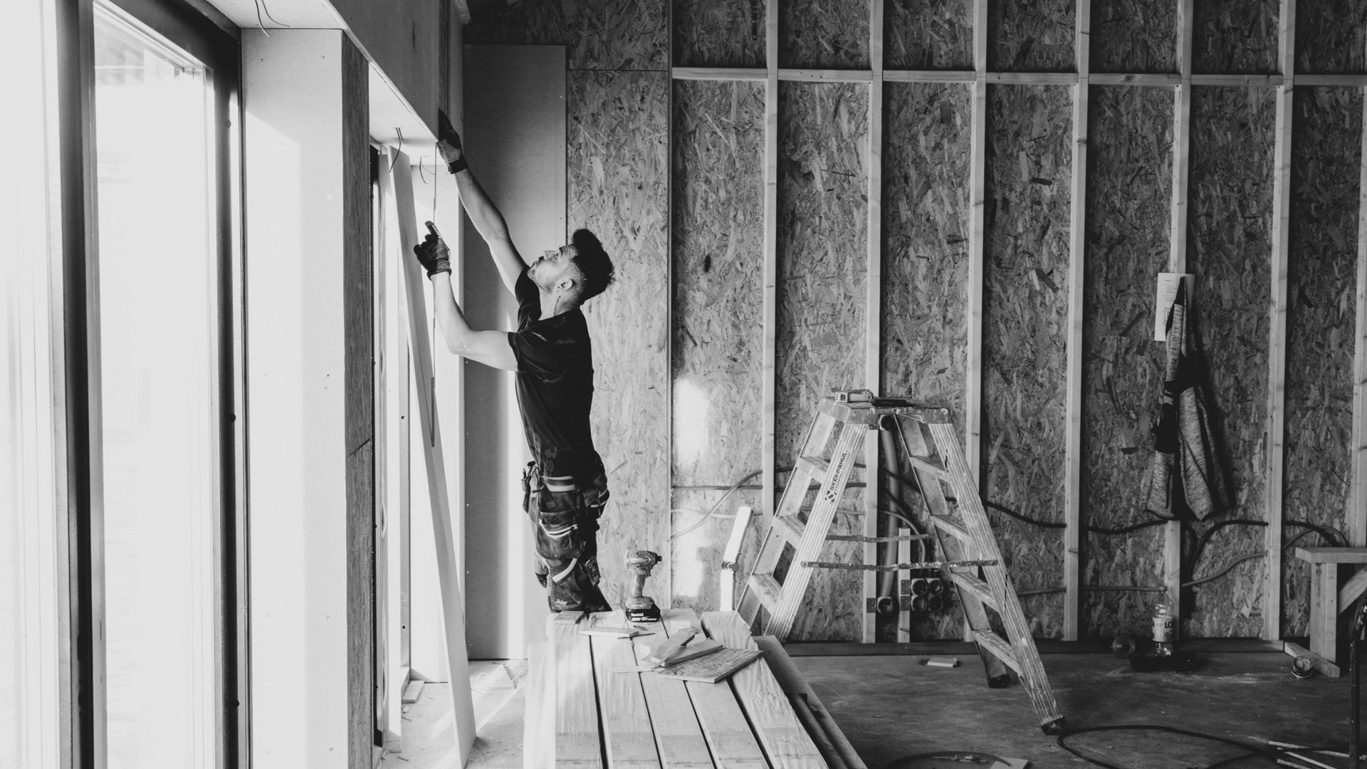 Worker installing drywall in an unfinished room with ladder and exposed framing