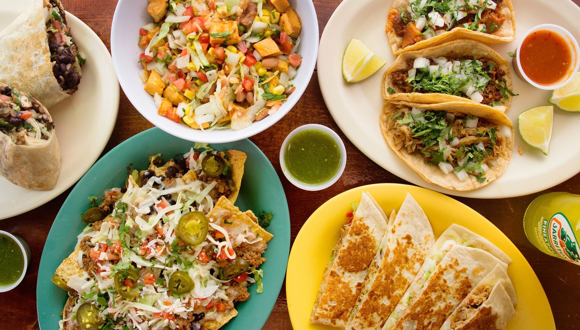 Various Mexican dishes: burritos, tacos, nachos, quesadillas, and a salad, on a wooden table with lime wedges.