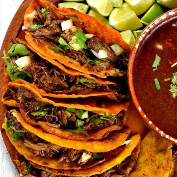 Birria tacos on a wooden platter with consome, lime wedges, and cilantro.