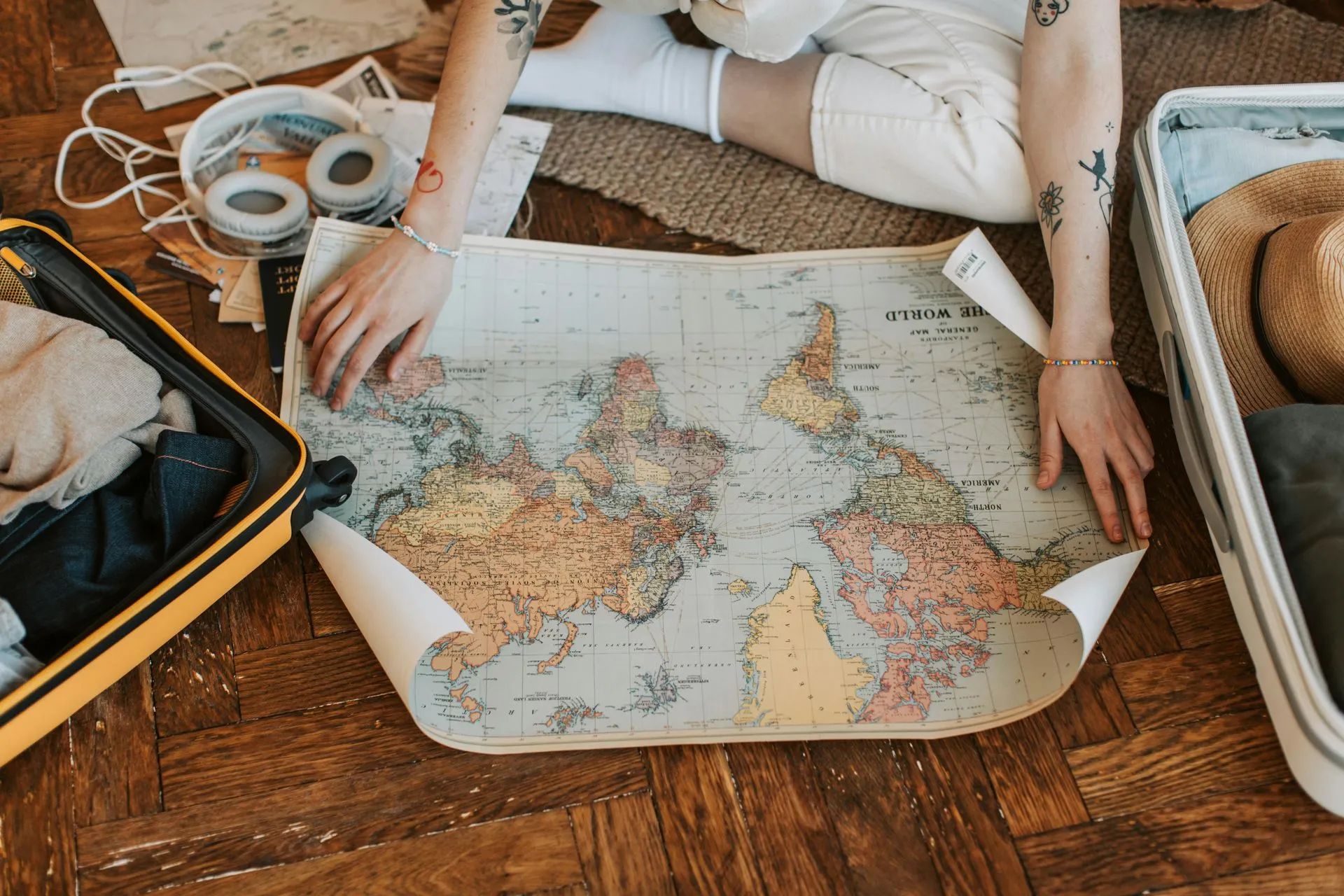 Person on floor with world map, packing suitcases; headphones and straw hat nearby.