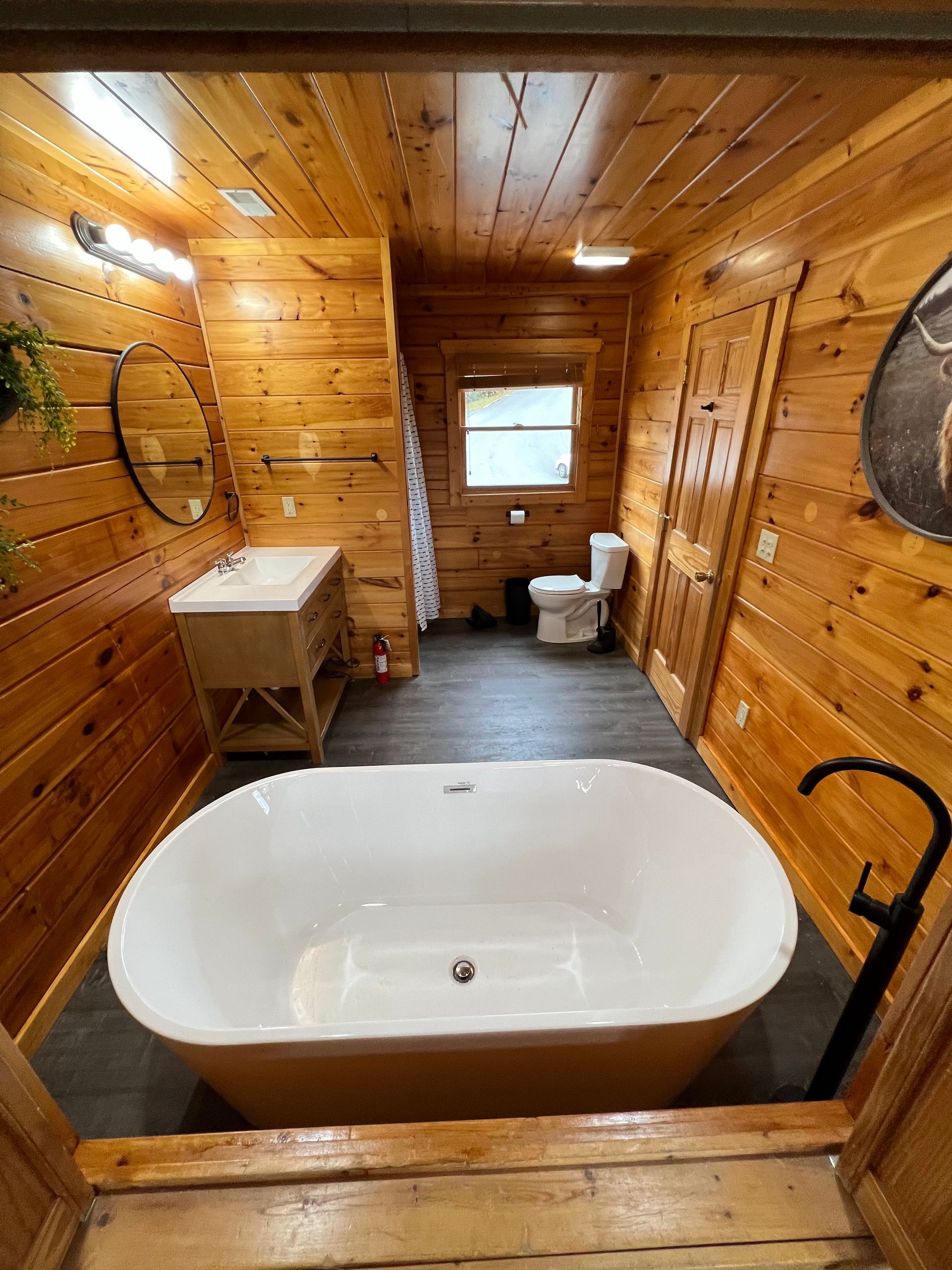 Rustic wood-paneled bathroom with a freestanding white tub, sink, and toilet under a small window.