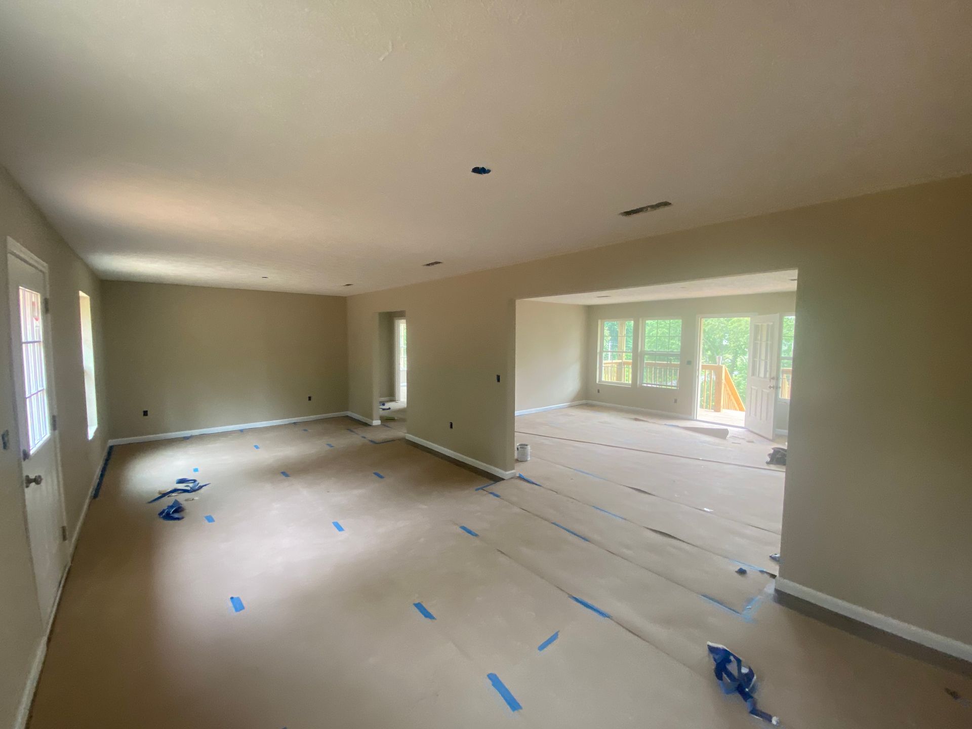 Empty beige living room under renovation, with taped floor coverings and large windows letting in daylight.