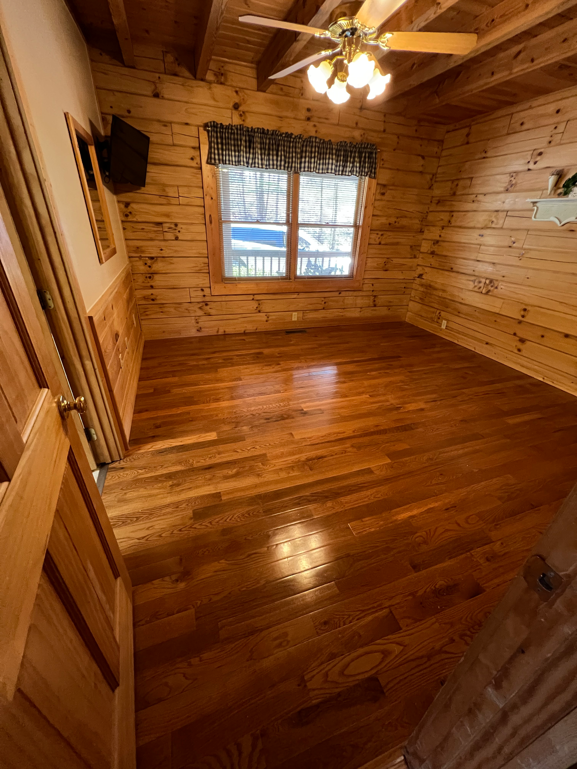 Empty rustic wooden room with polished floors, log walls, ceiling fan, and a window with curtains.