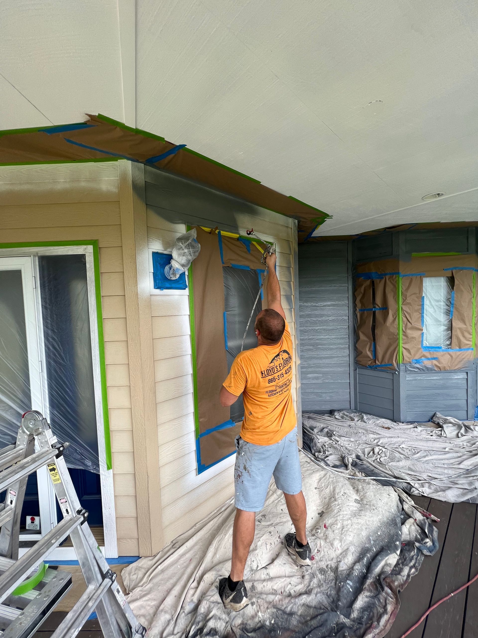 Person painting exterior trim on a house under renovation, standing on tarp-covered ground beside ladders.