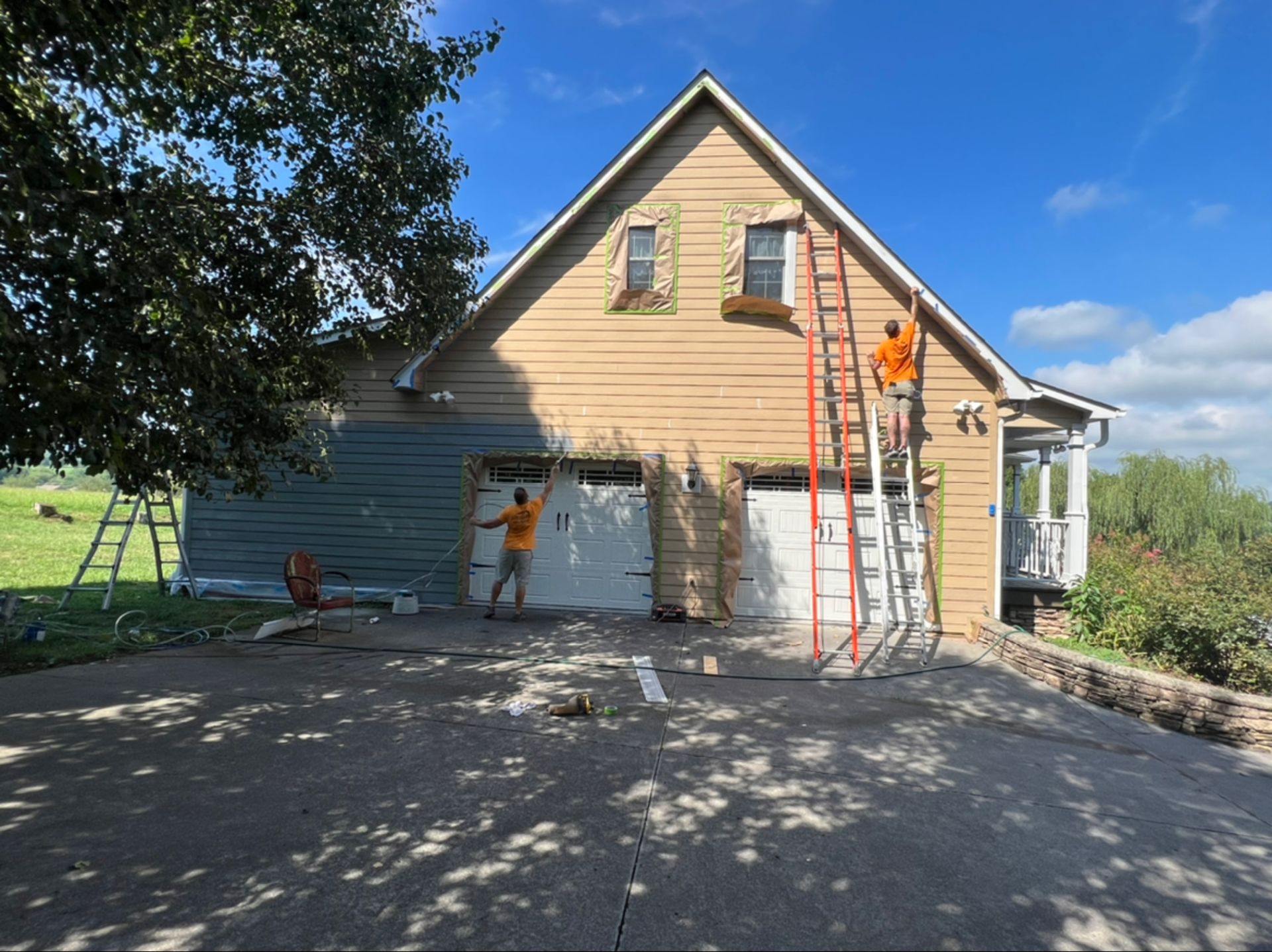 Workers painting the exterior of a tan house with ladders on a sunny day