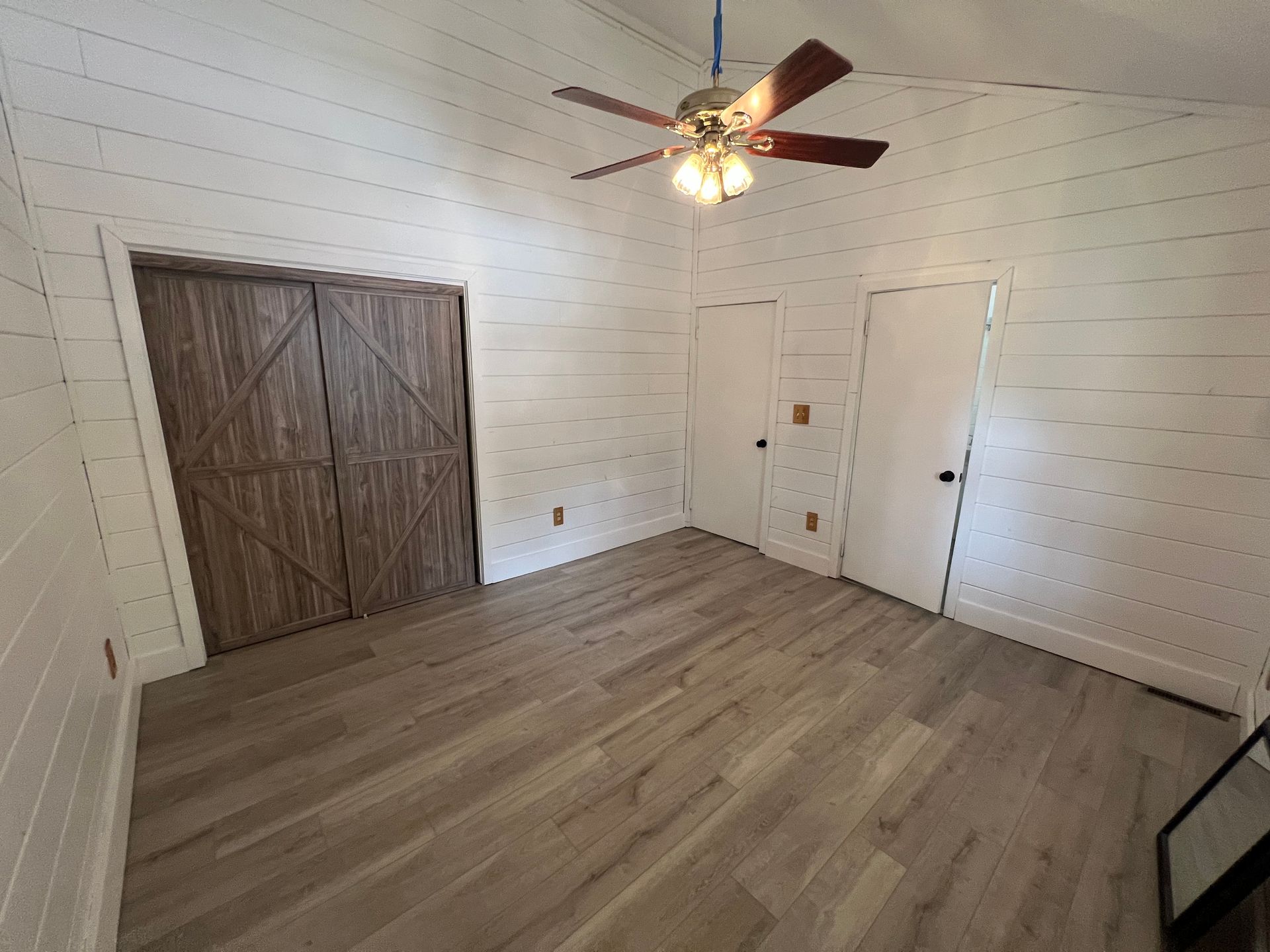 Empty white bedroom with wood-look floor, ceiling fan, double closet doors, and a single door.