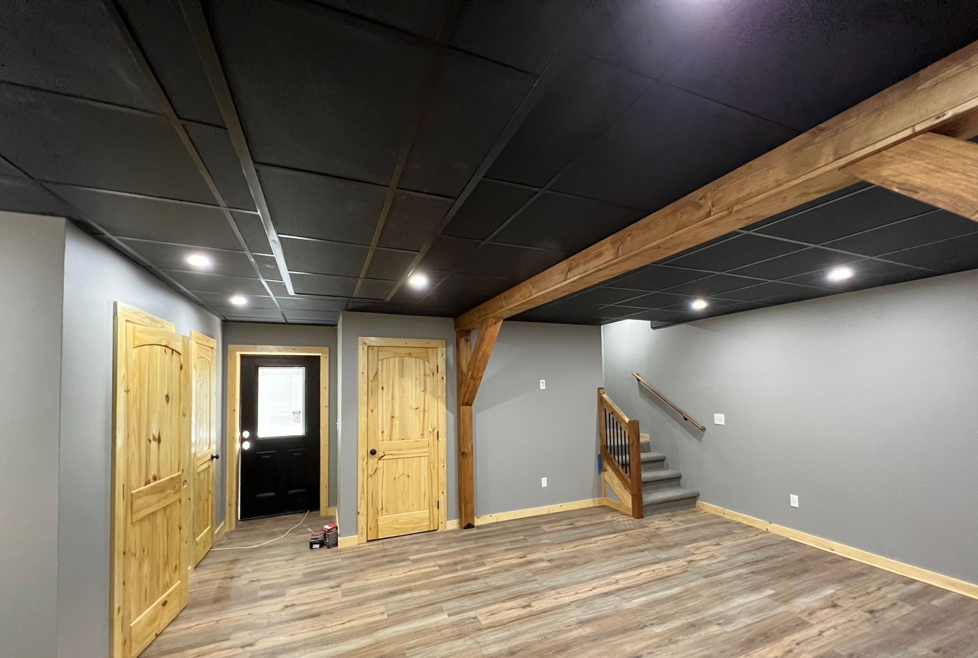 Empty finished basement with gray walls, wood floors, black ceiling, wooden doors, and a small staircase.