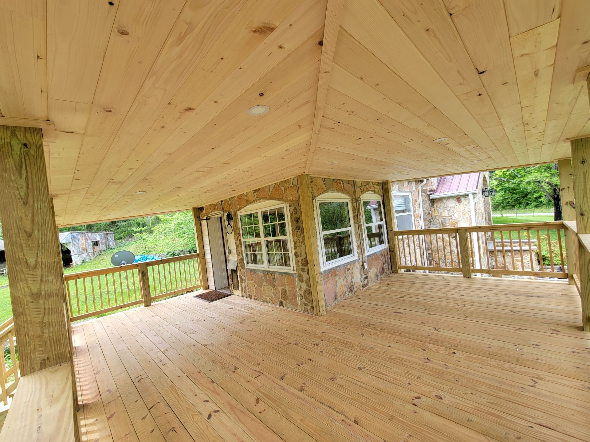 Wooden covered porch with wraparound railing and exposed ceiling, overlooking a green yard.