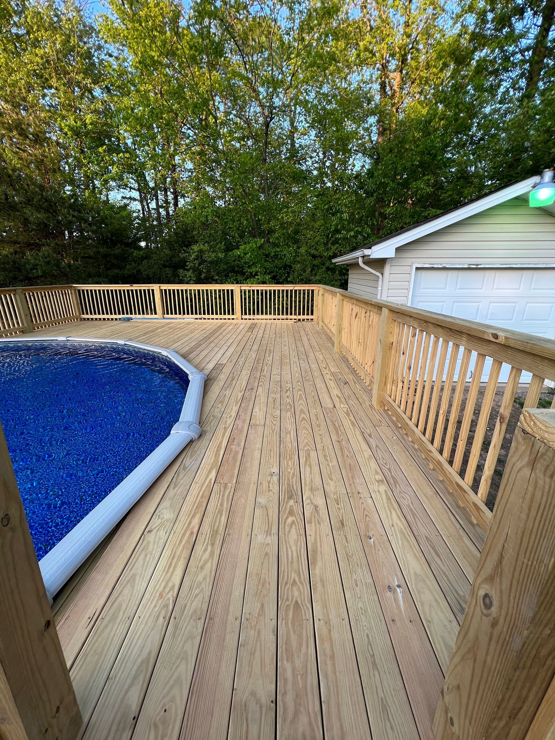 Wooden deck beside a blue above-ground pool, lined with railings and surrounded by trees.