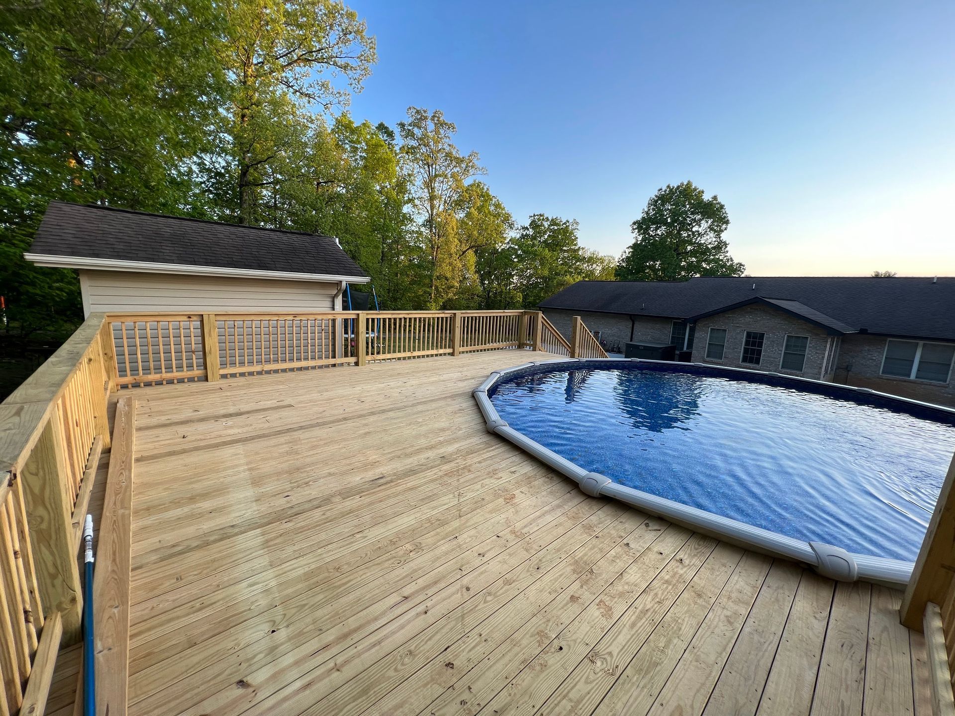 Wooden deck curving around a blue swimming pool beside a house, with trees and a clear sky.