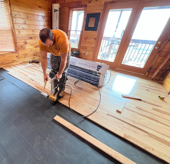 Worker installing a wood floor inside a sunlit room with large windows