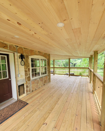 Covered wooden porch with knotty pine ceiling, stone wall, windows, and railing overlooking greenery