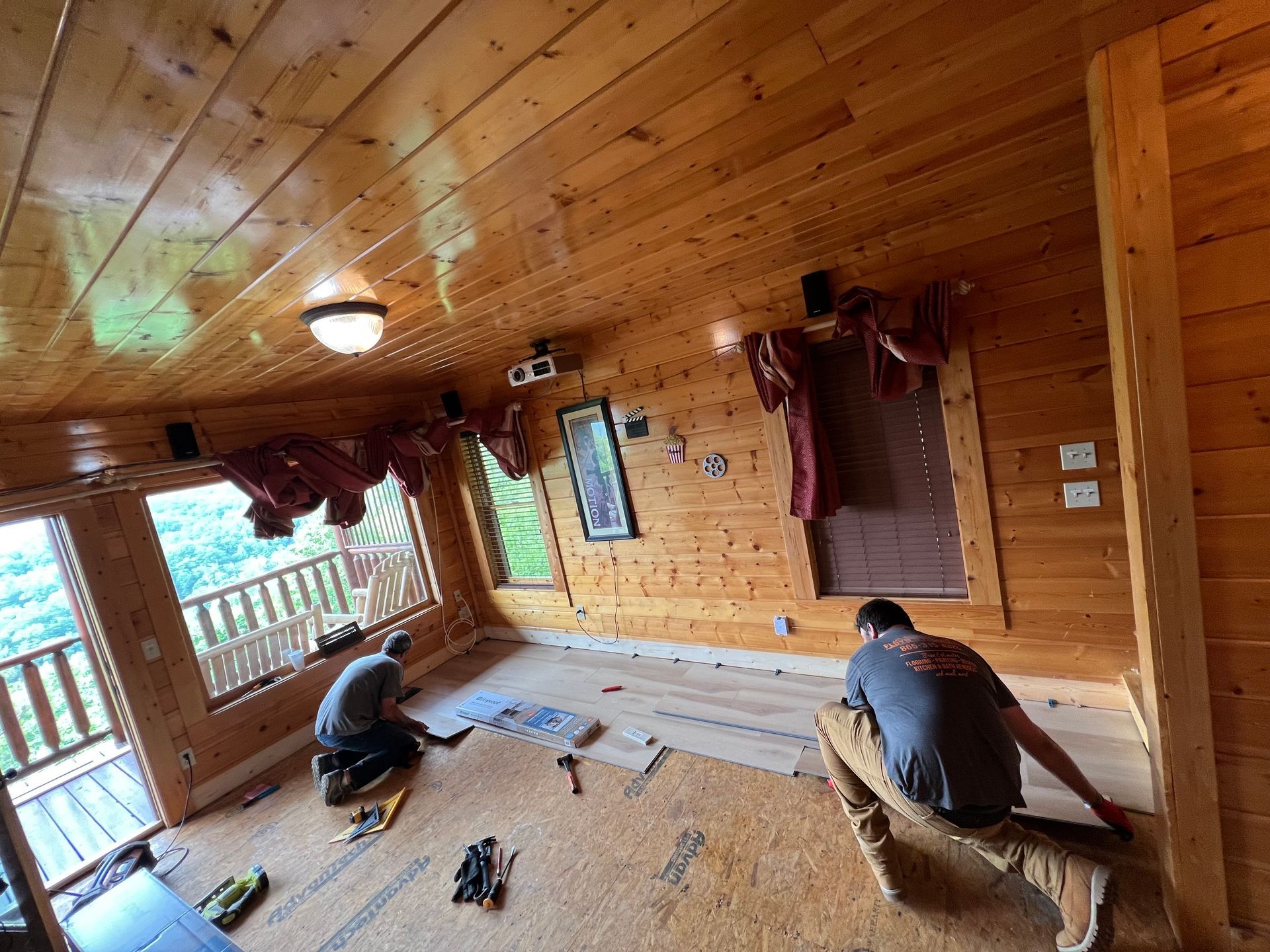 Two workers on a wooden porch installing floor planks in a cabin-like room.