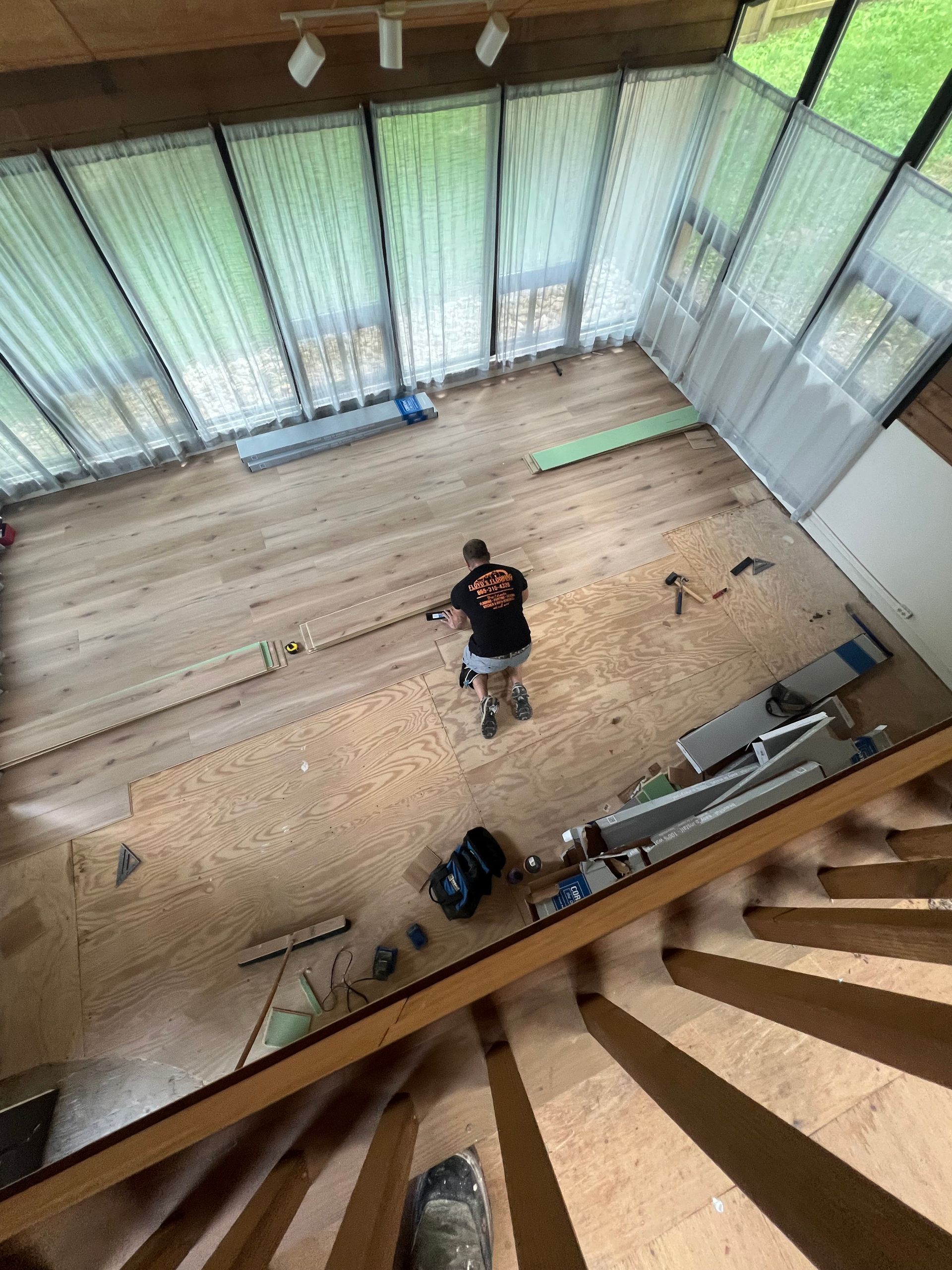 High-angle view of a person standing in a bright, wood-floored room with large windows and scattered tools.