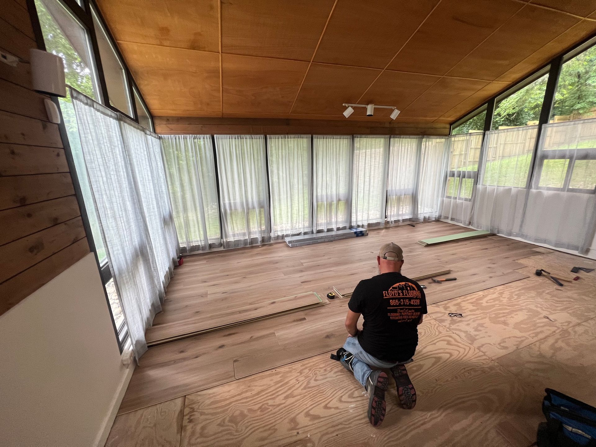 Person kneeling on a bare floor in a screened porch with curtains and a wood ceiling