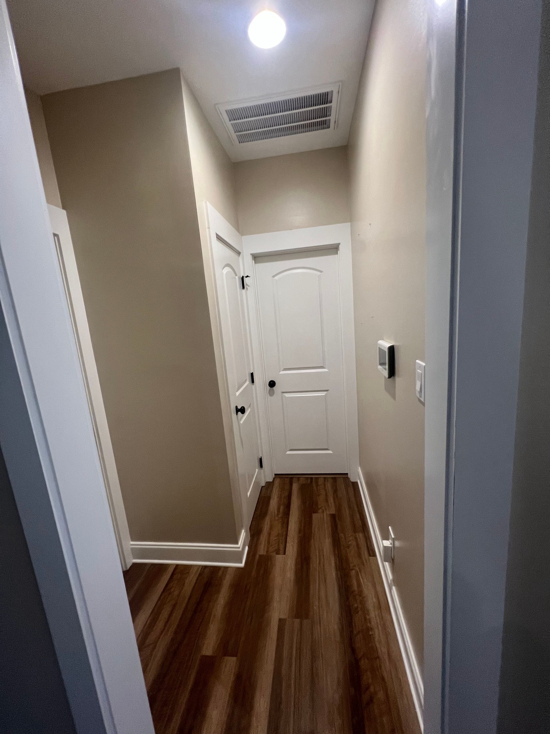 Narrow hallway with wood floors, beige walls, white doors, and a ceiling light.