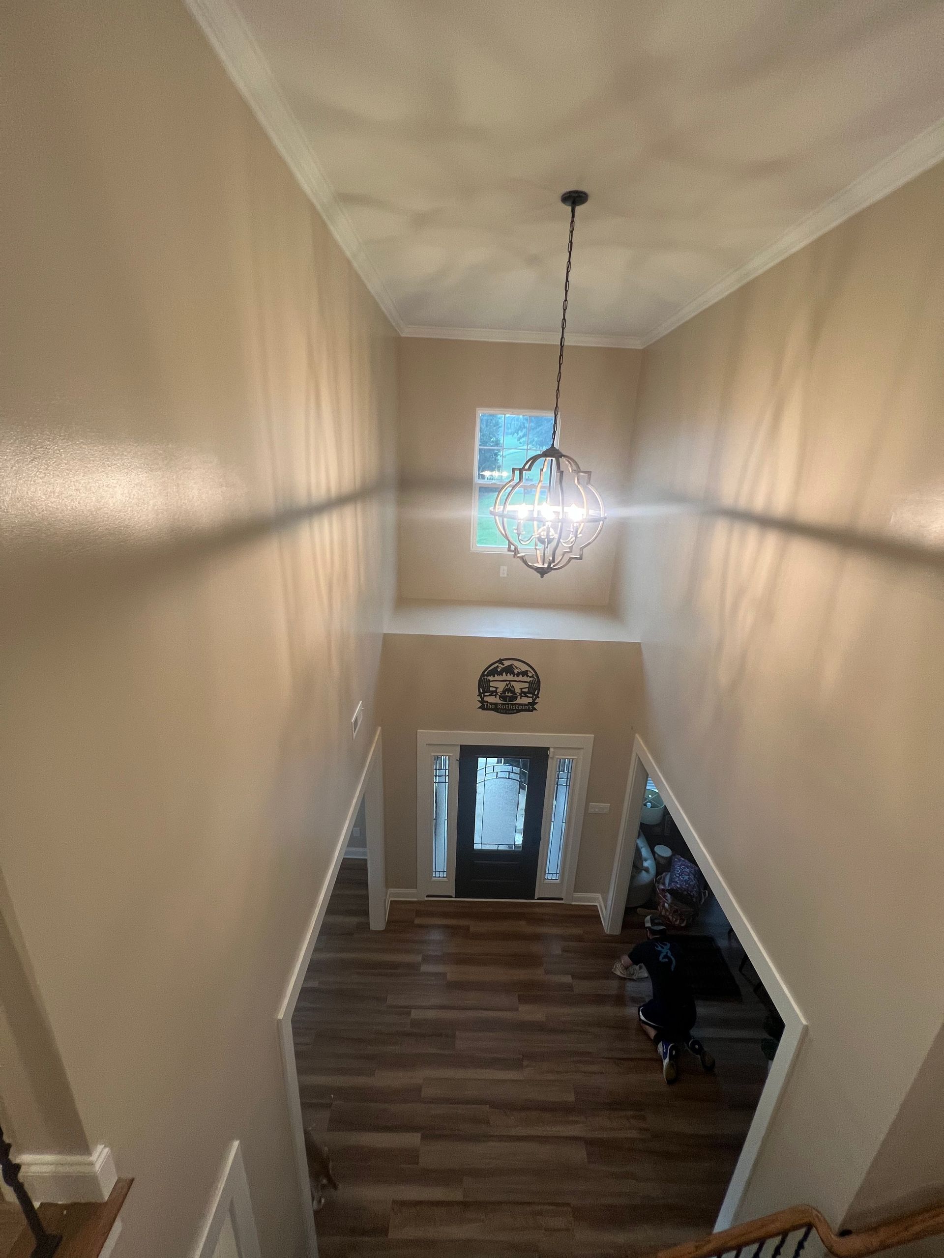 View down a narrow stairwell with wood floors, beige walls, and a hanging light toward a front door and window.