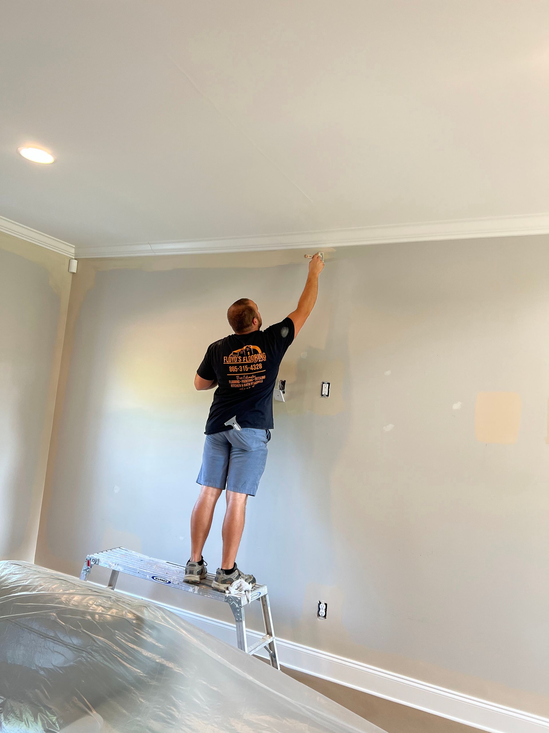 Person painting a wall in a sparsely furnished room, standing on a covered ladder.