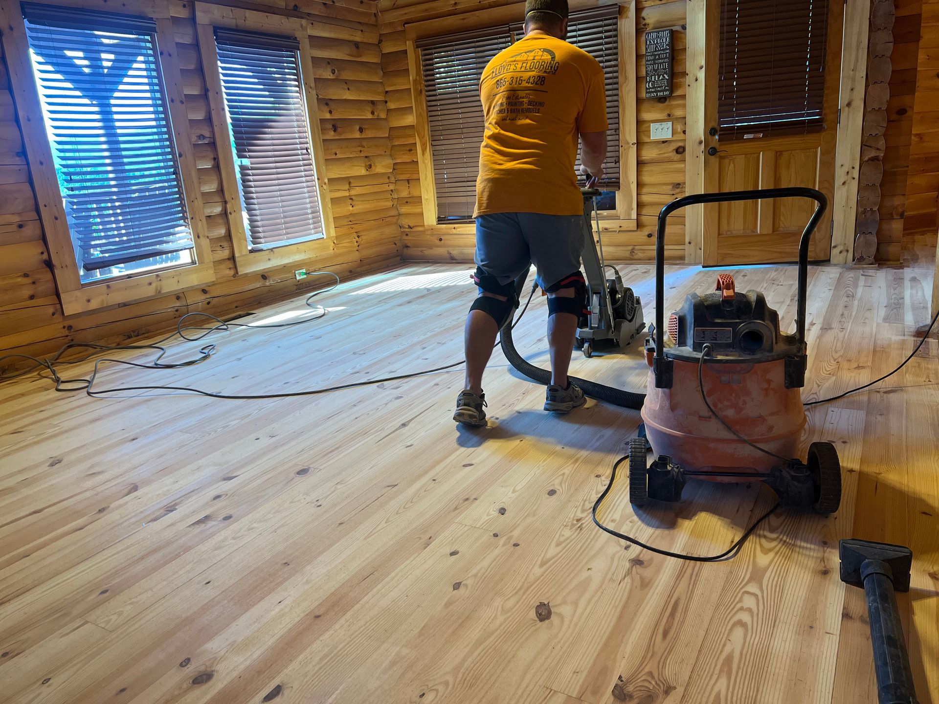 Person sanding a wooden floor in an unfinished room with exposed brick walls and windows.