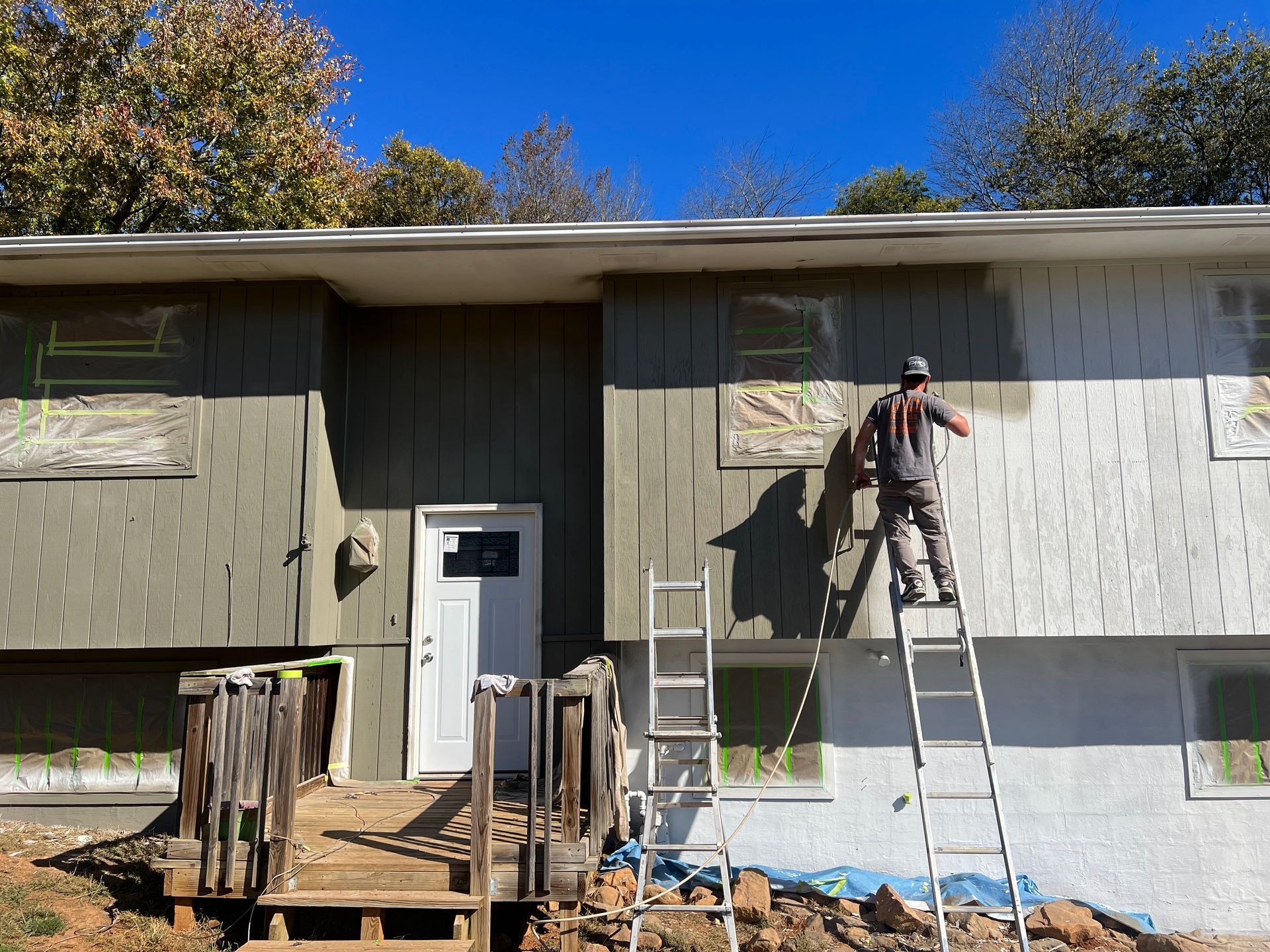 Worker on ladder painting gray siding on a house exterior under a clear blue sky