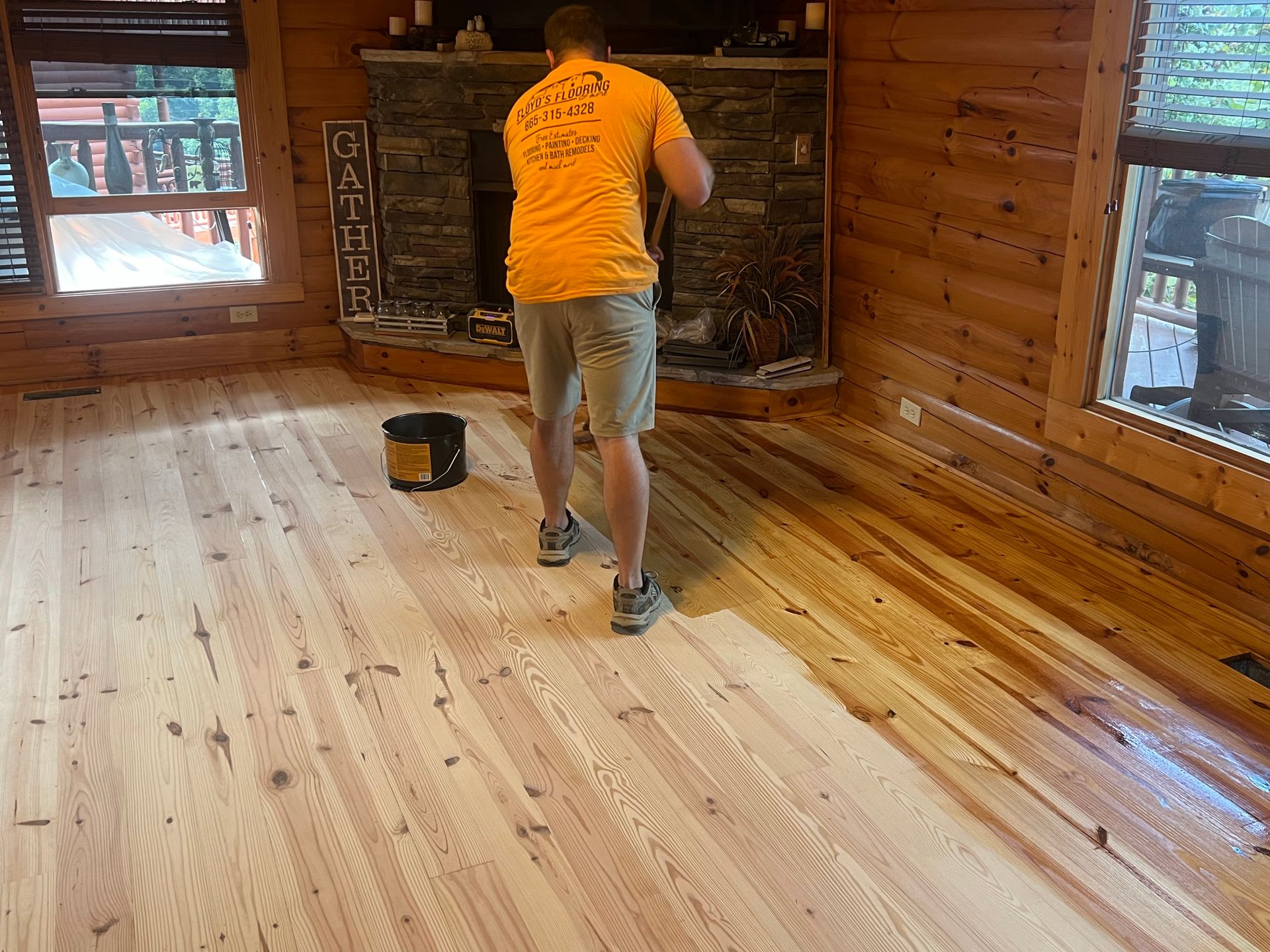 Person painting a wood-paneled room, with a bucket on the floor and windows on both sides