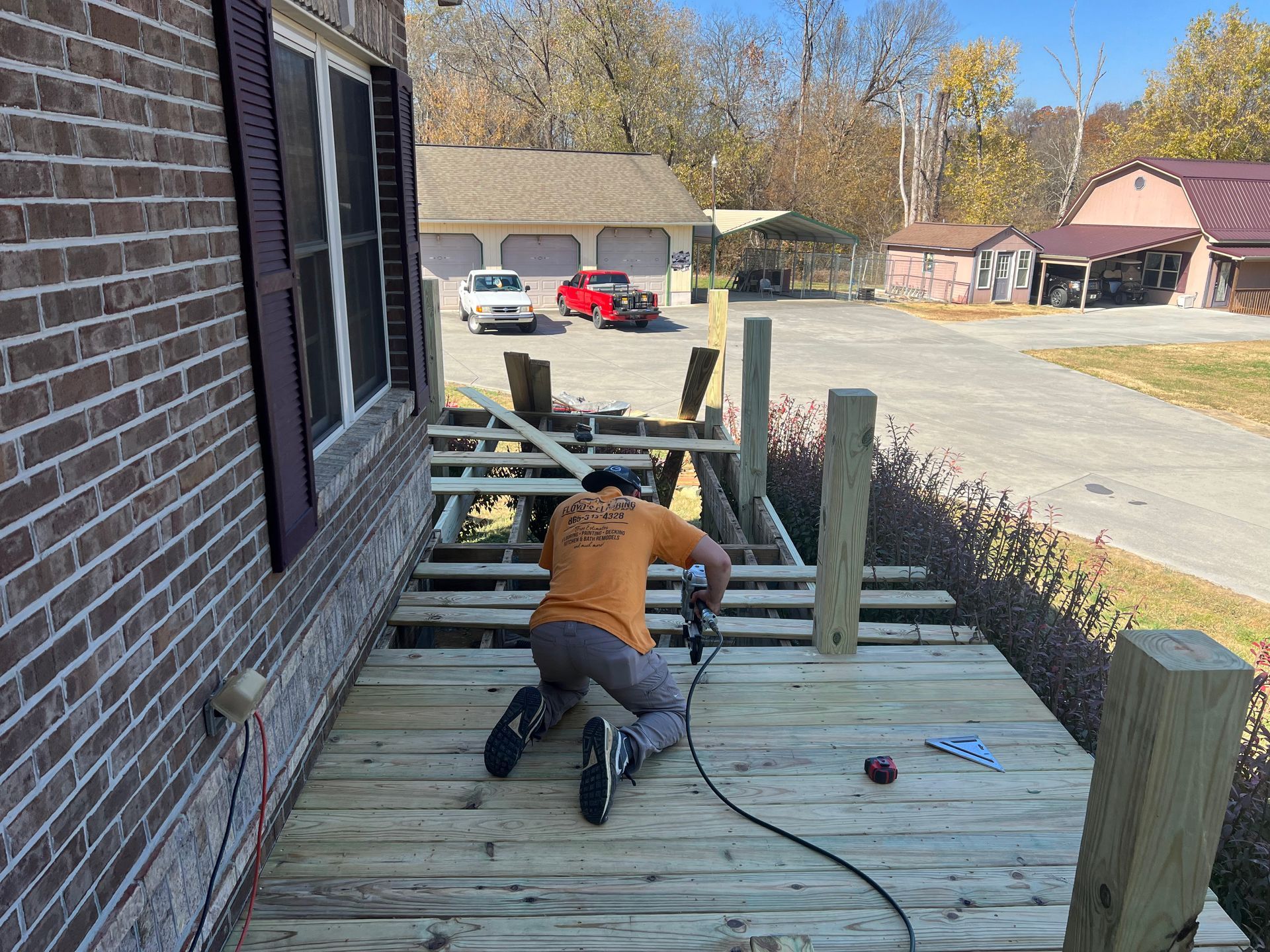 Worker installing wooden deck boards beside a brick house, with posts and tools on the deck.