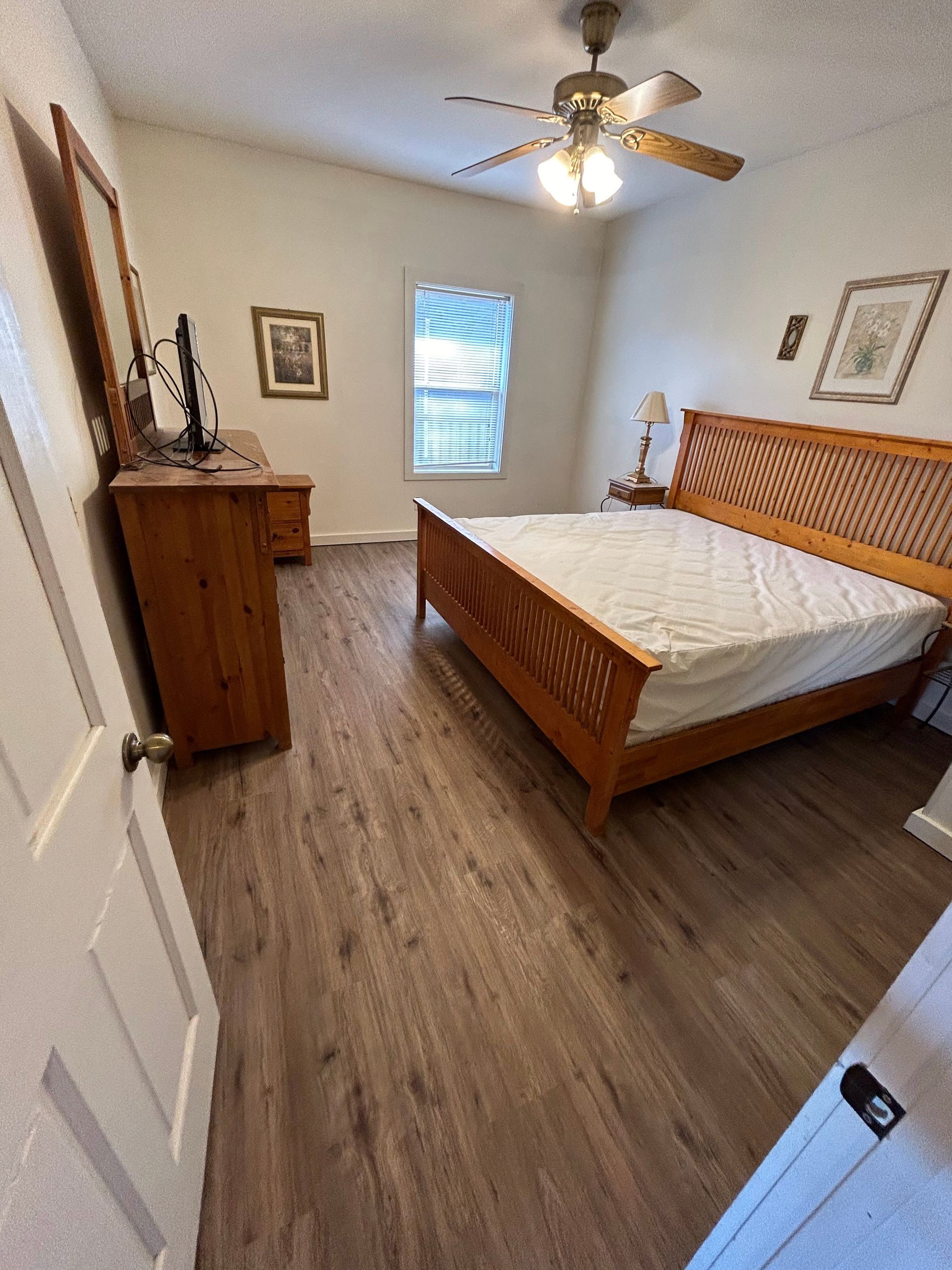 Bright bedroom with a wooden bed, dresser, ceiling fan, and window on wood flooring.