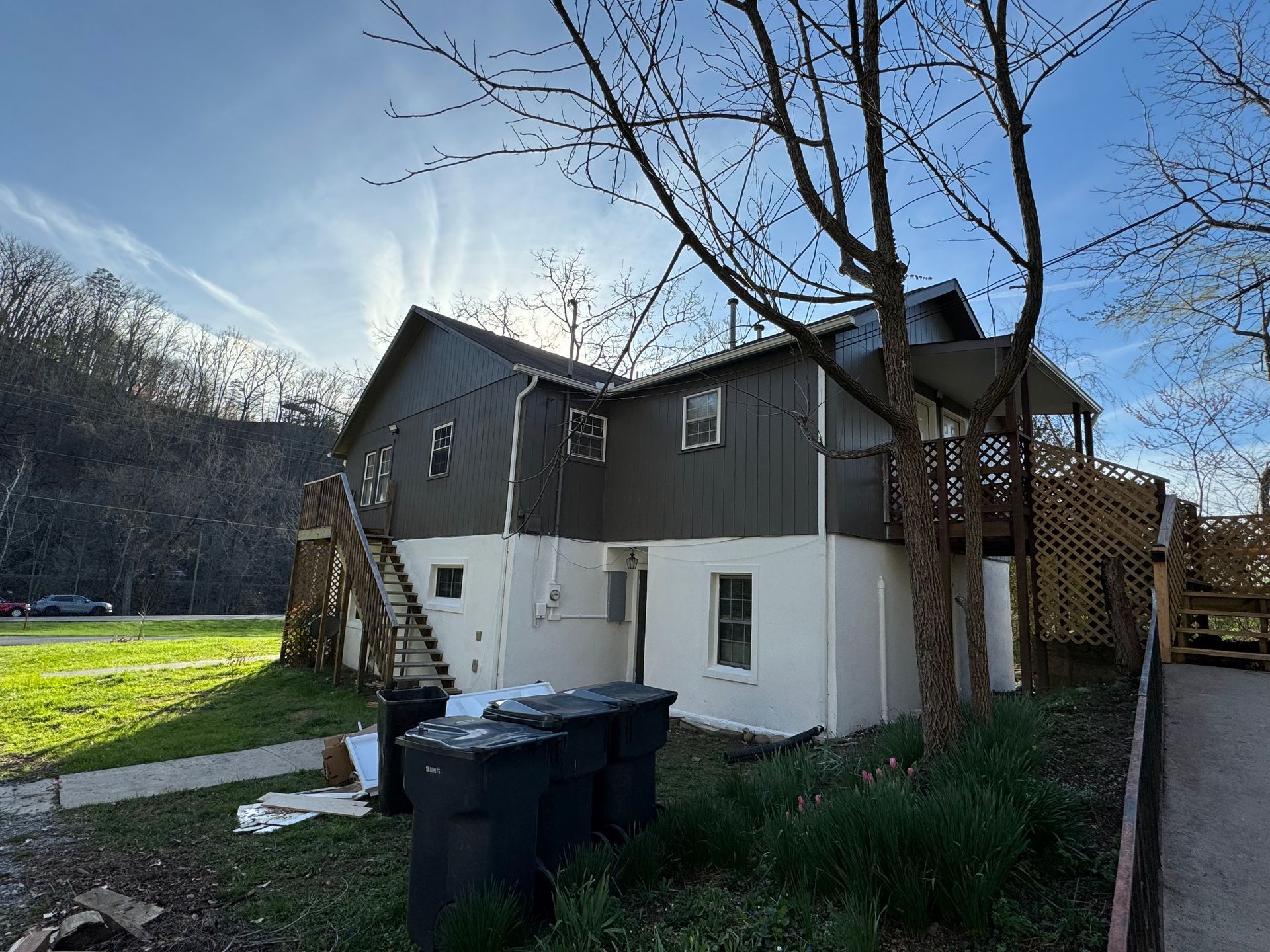 Two-story house with dark gray and white siding, trees, and trash bins in the yard beside a walkway.