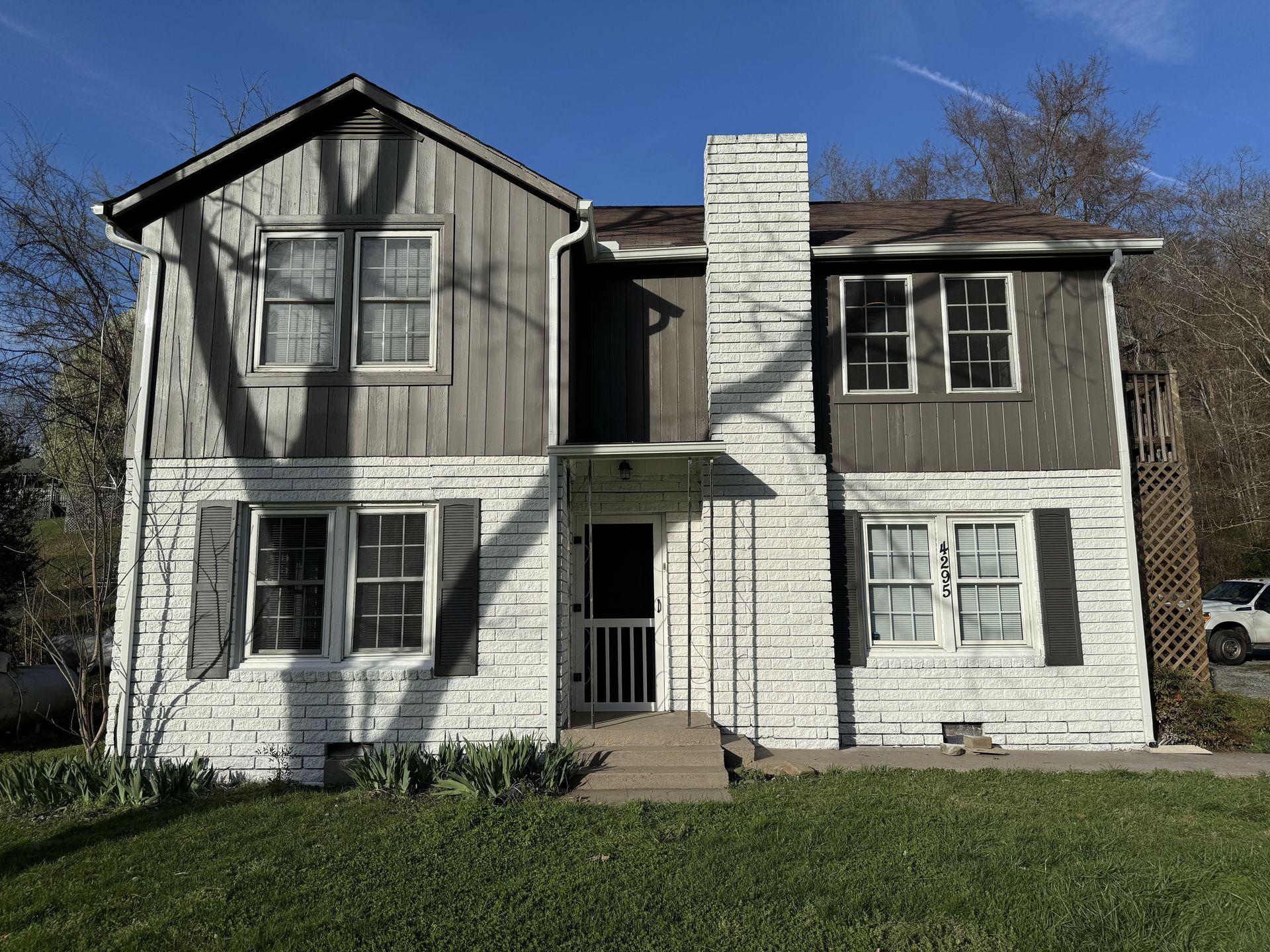 Two-story house with white stone and weathered wood siding, central chimney, and a small front lawn.