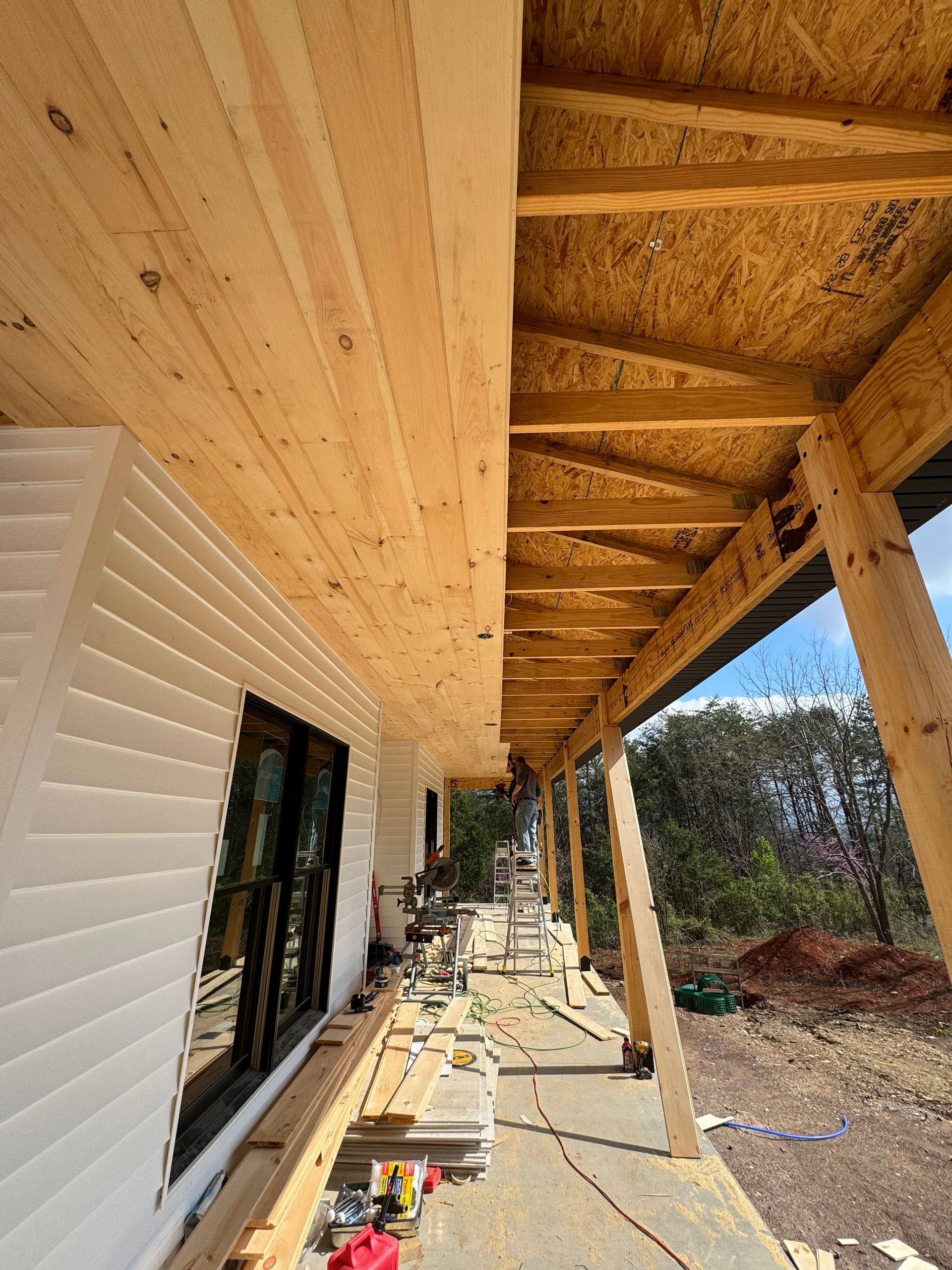 Covered porch with exposed wooden rafters beside a white house, construction materials scattered along the walkway.