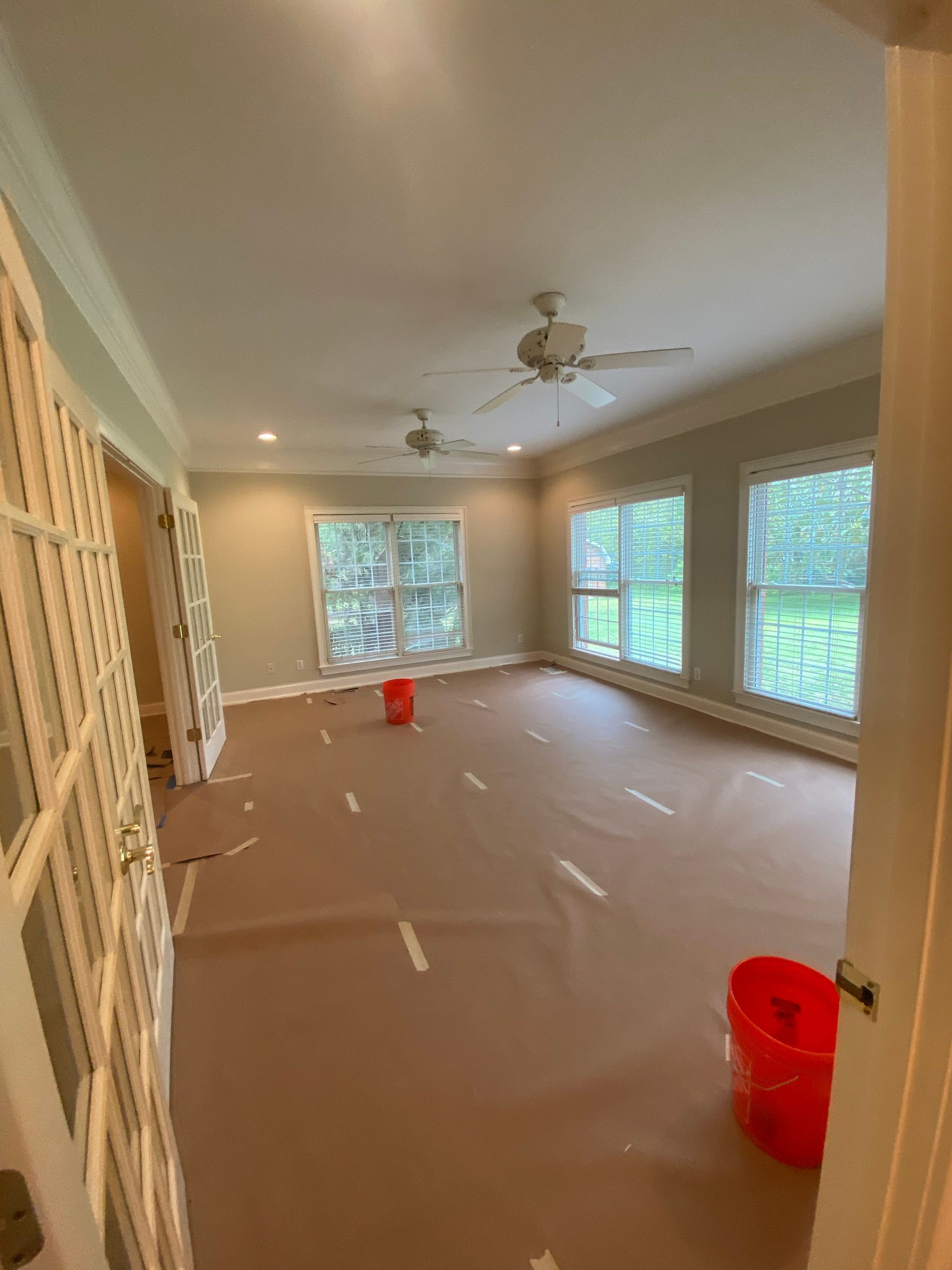 Empty sunroom with beige carpet, ceiling fan, and large windows, with a red bucket on the floor