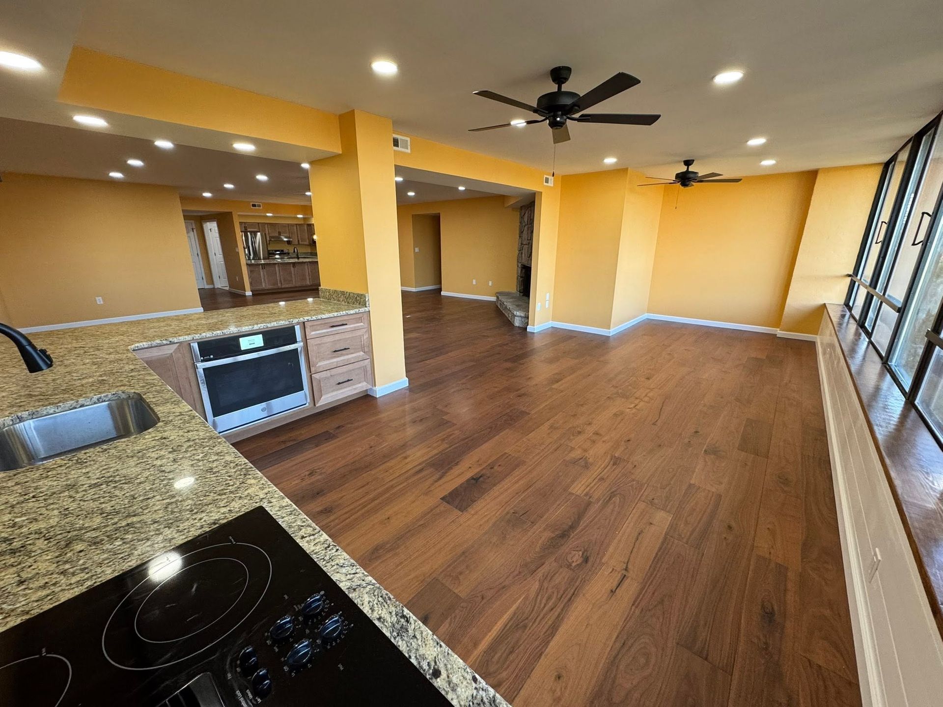 Empty open-plan kitchen and living area with wood floors, tan walls, ceiling fans, and large windows.