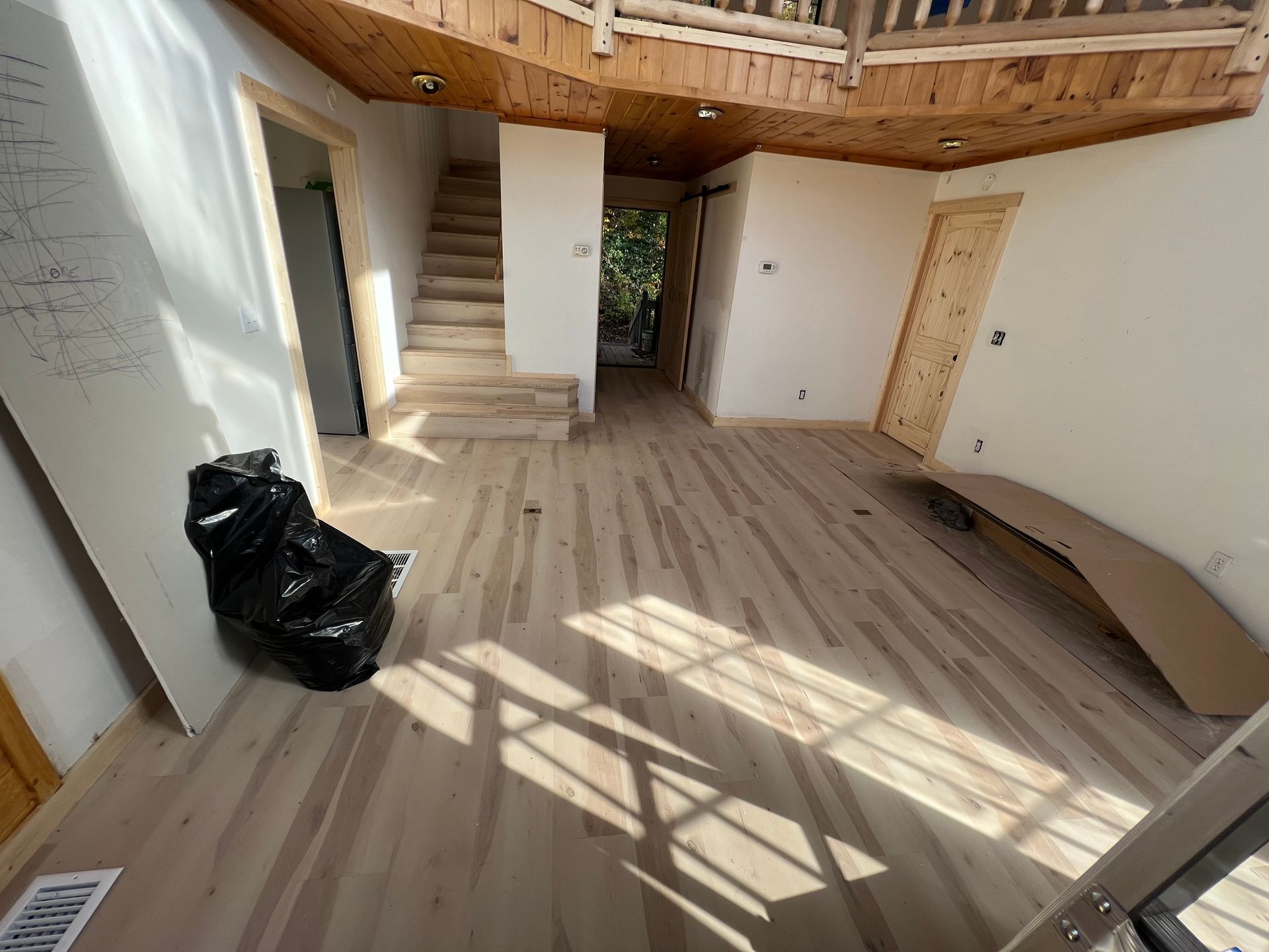 Empty unfinished room with wooden floor, stairs, loft railing, sunlight on floor, and a black trash bag by the wall