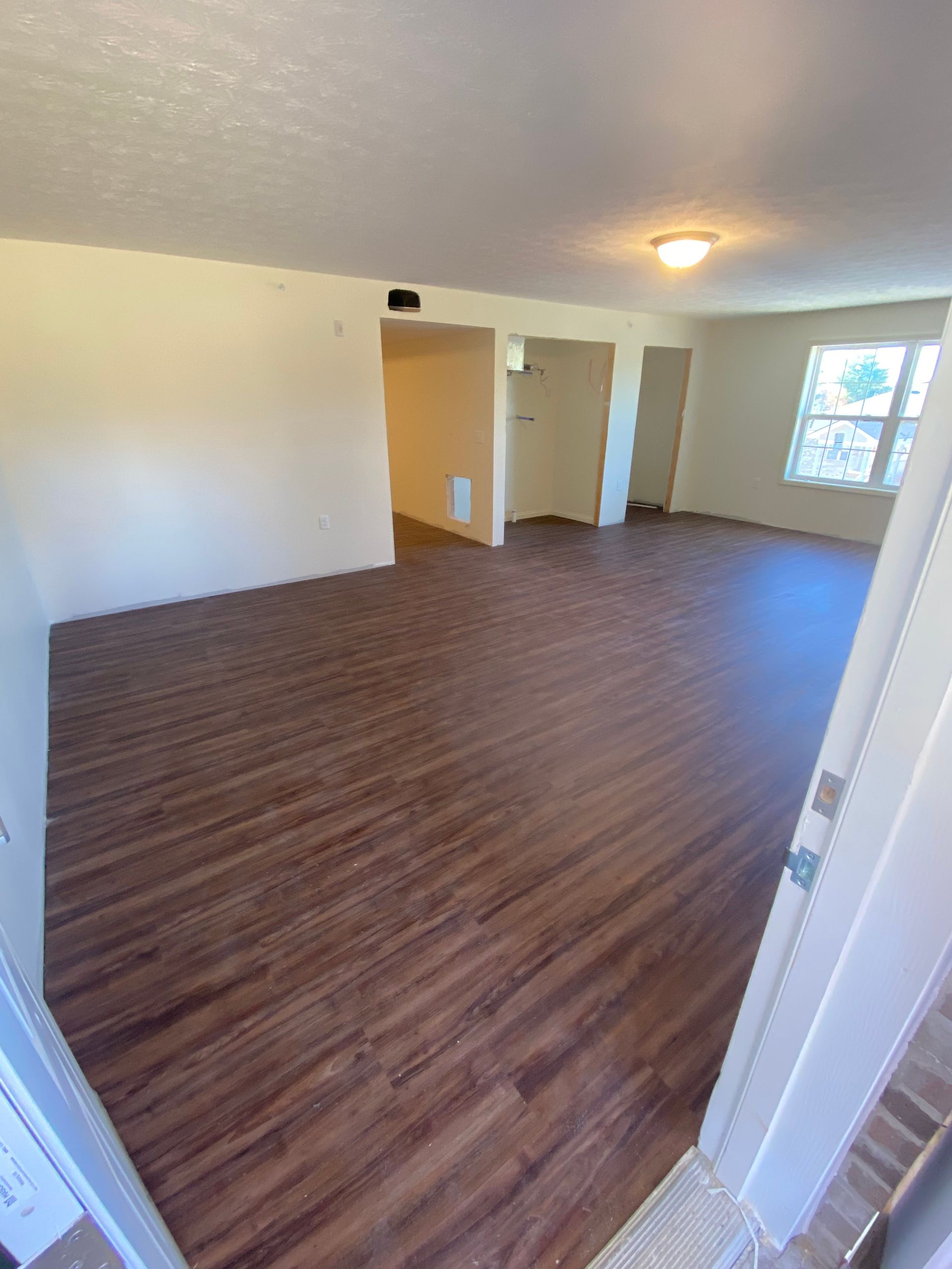 Empty living room with brown wood-look flooring, white walls, and a window on the far right.